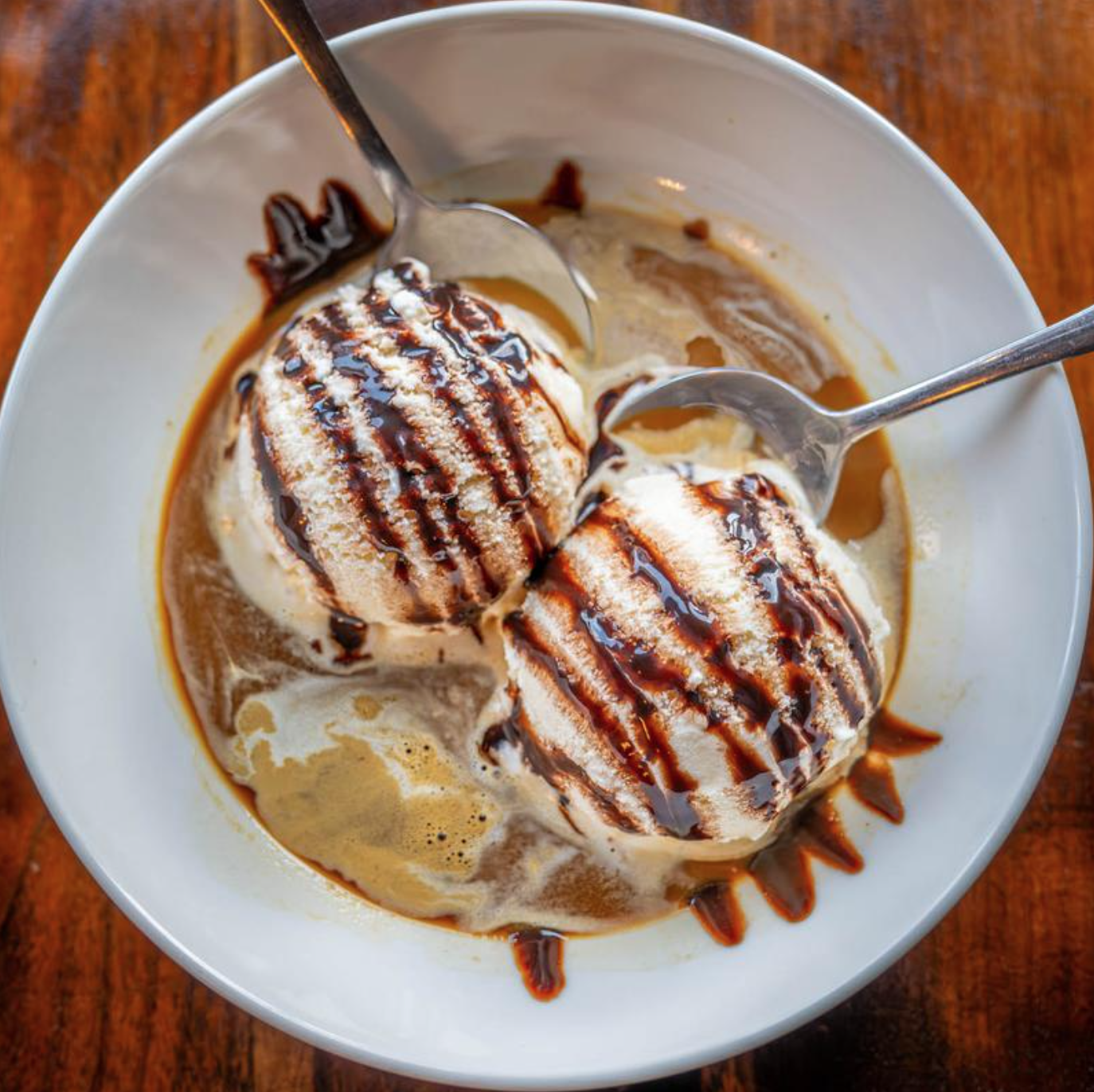 Two scoops of vanilla ice cream topped with chocolate syrup in a white bowl on a wooden table.