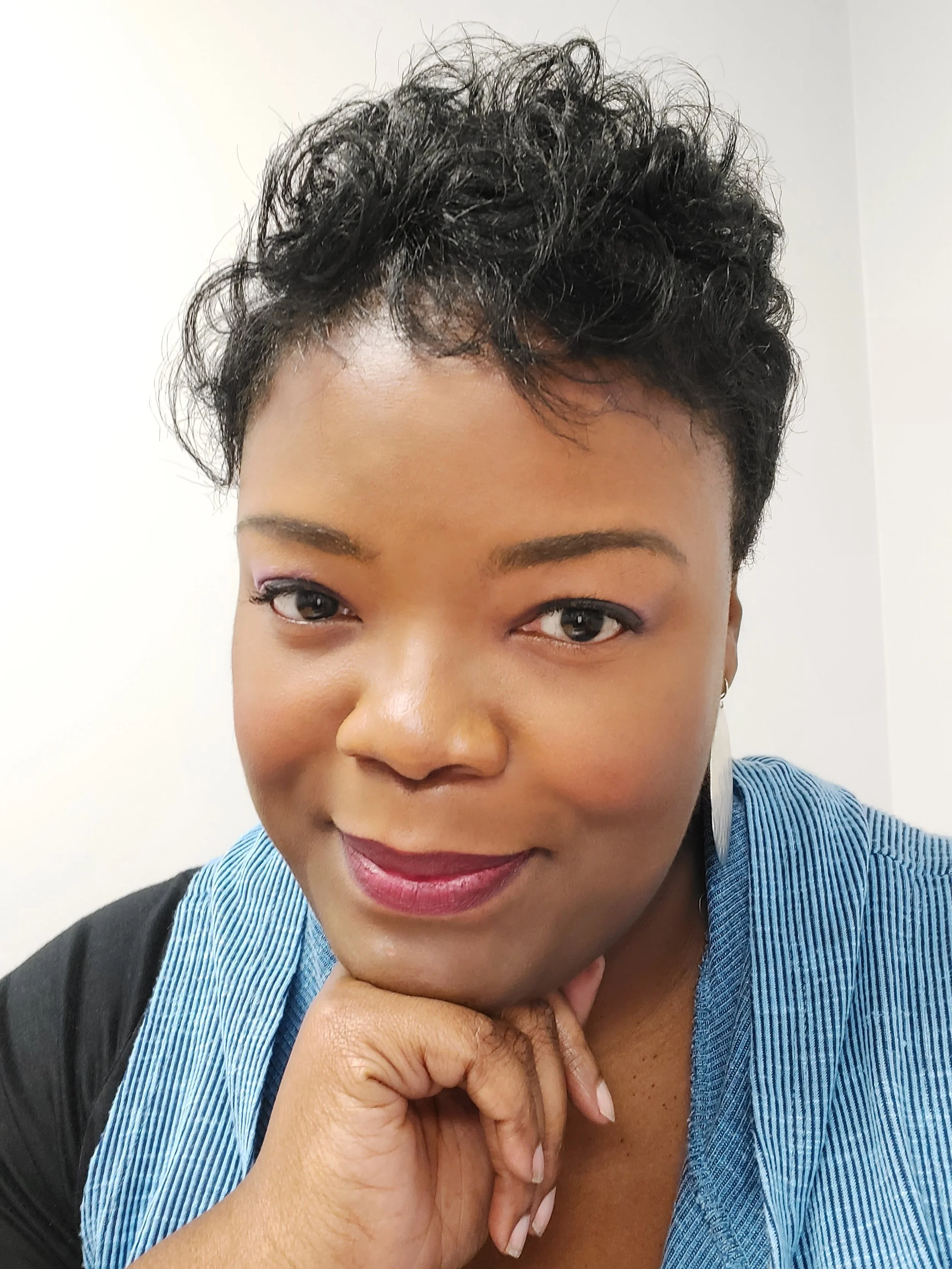 Close-up of a smiling woman with short, curly black hair, wearing a blue striped shirt and light makeup, resting her chin on her hand.