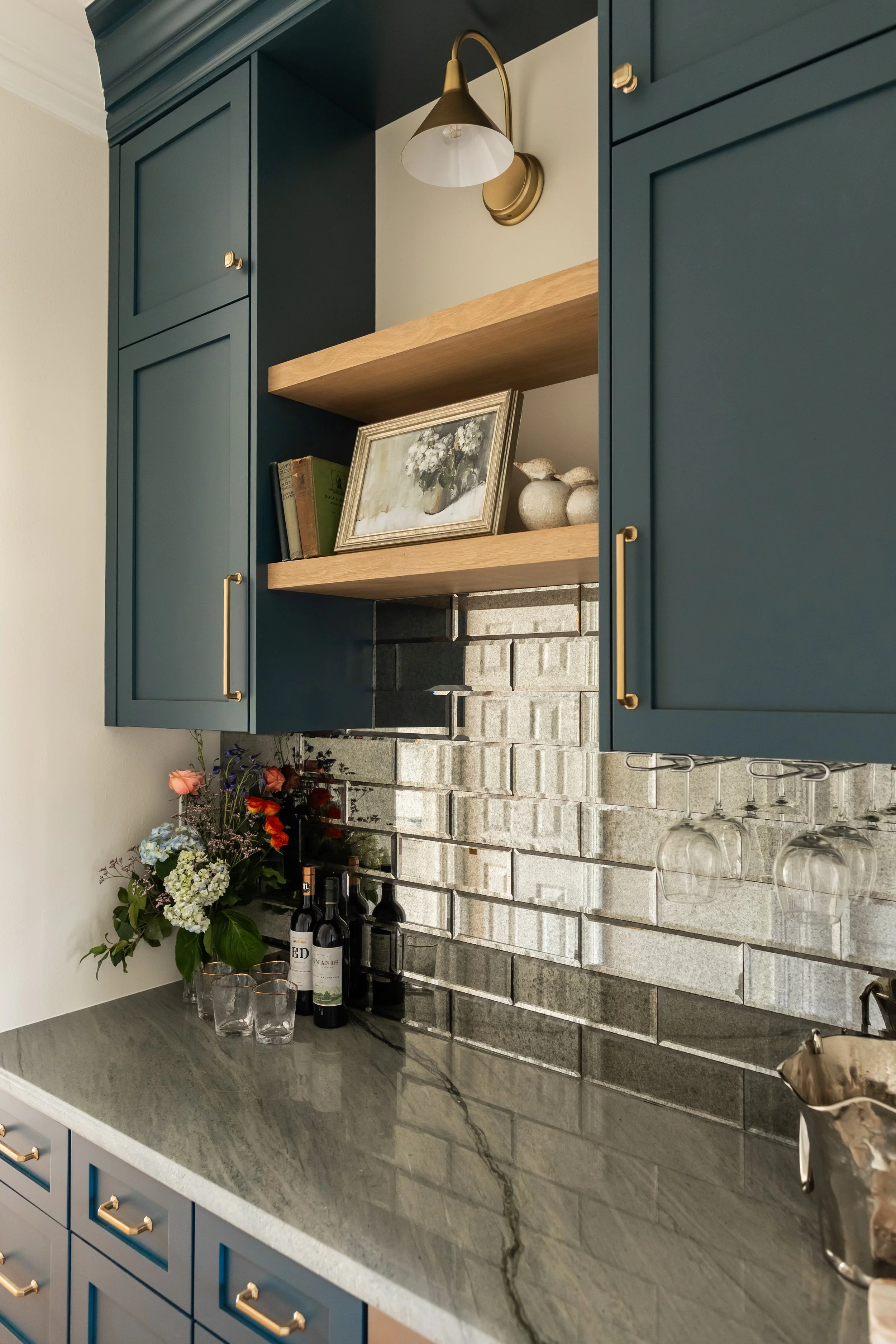 This custom bar creates a moody yet refined moment, anchored by deep blue shaker cabinetry and warm brass hardware that add richness and contrast. A dimensional stone tile backsplash brings texture and depth, while the open wood shelf softens the pal