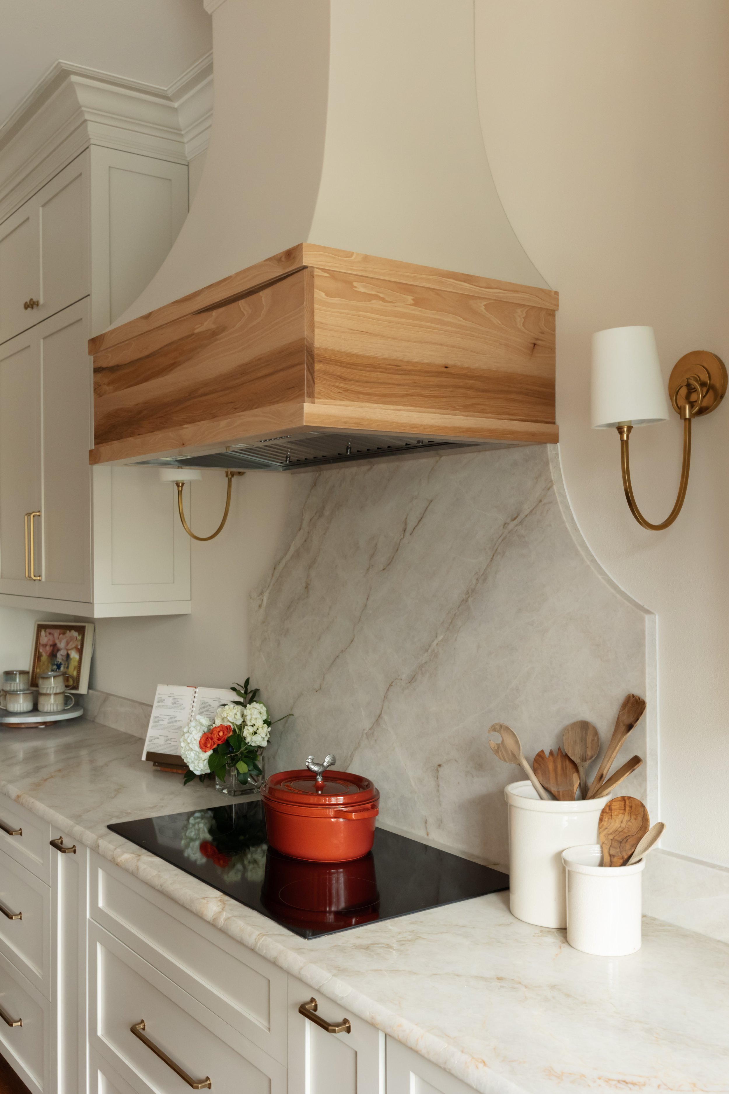 The kitchen stove is centered beneath a custom wood range hood, creating a natural focal point along the back wall. A marble backsplash with soft gray veining adds elegance and contrast while keeping the palette refined. Sculptural pendant lights wit