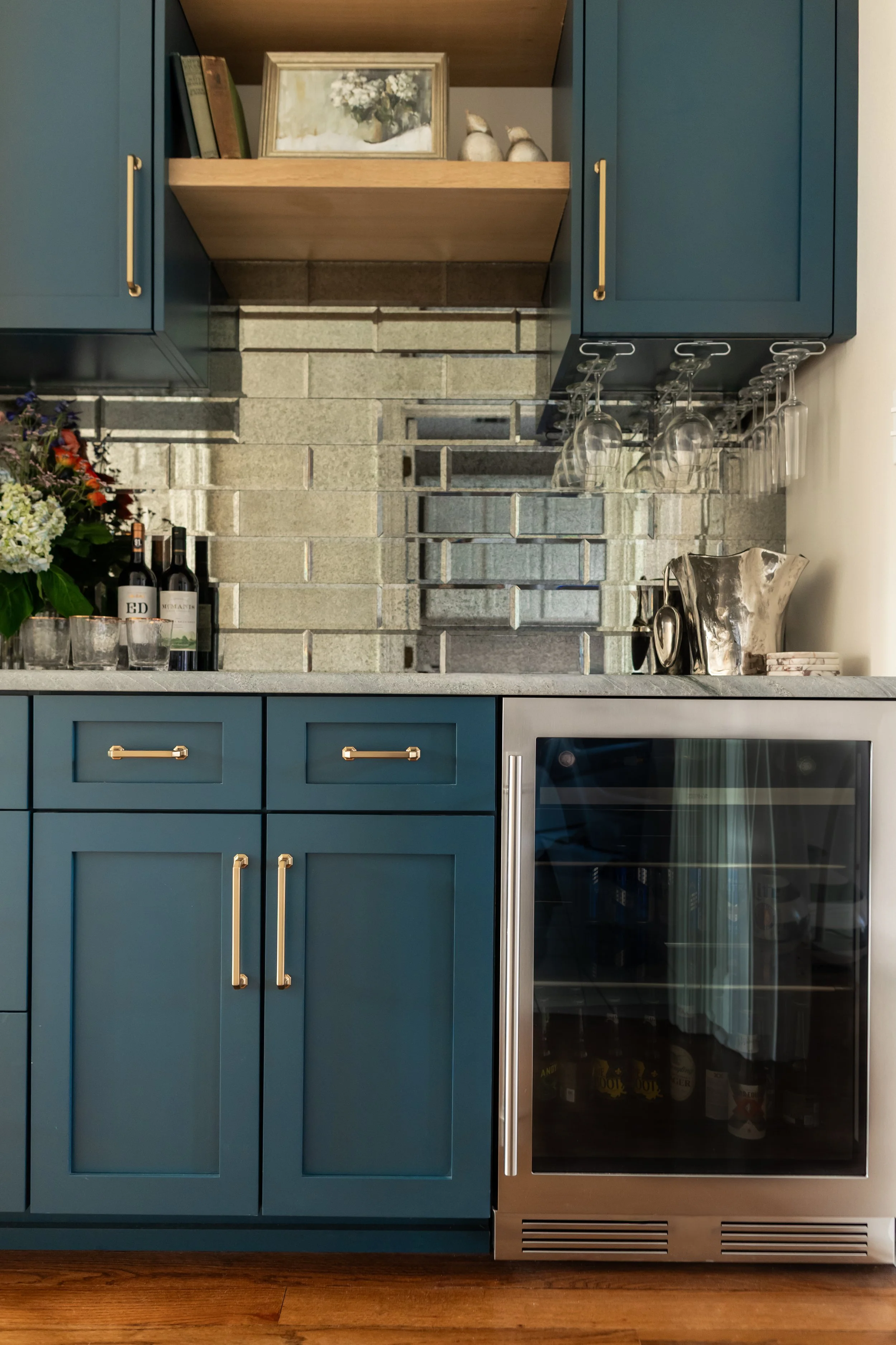 This custom bar creates a moody yet refined moment, anchored by deep blue shaker cabinetry and warm brass hardware that add richness and contrast. A dimensional stone tile backsplash brings texture and depth, while the open wood shelf softens the pal
