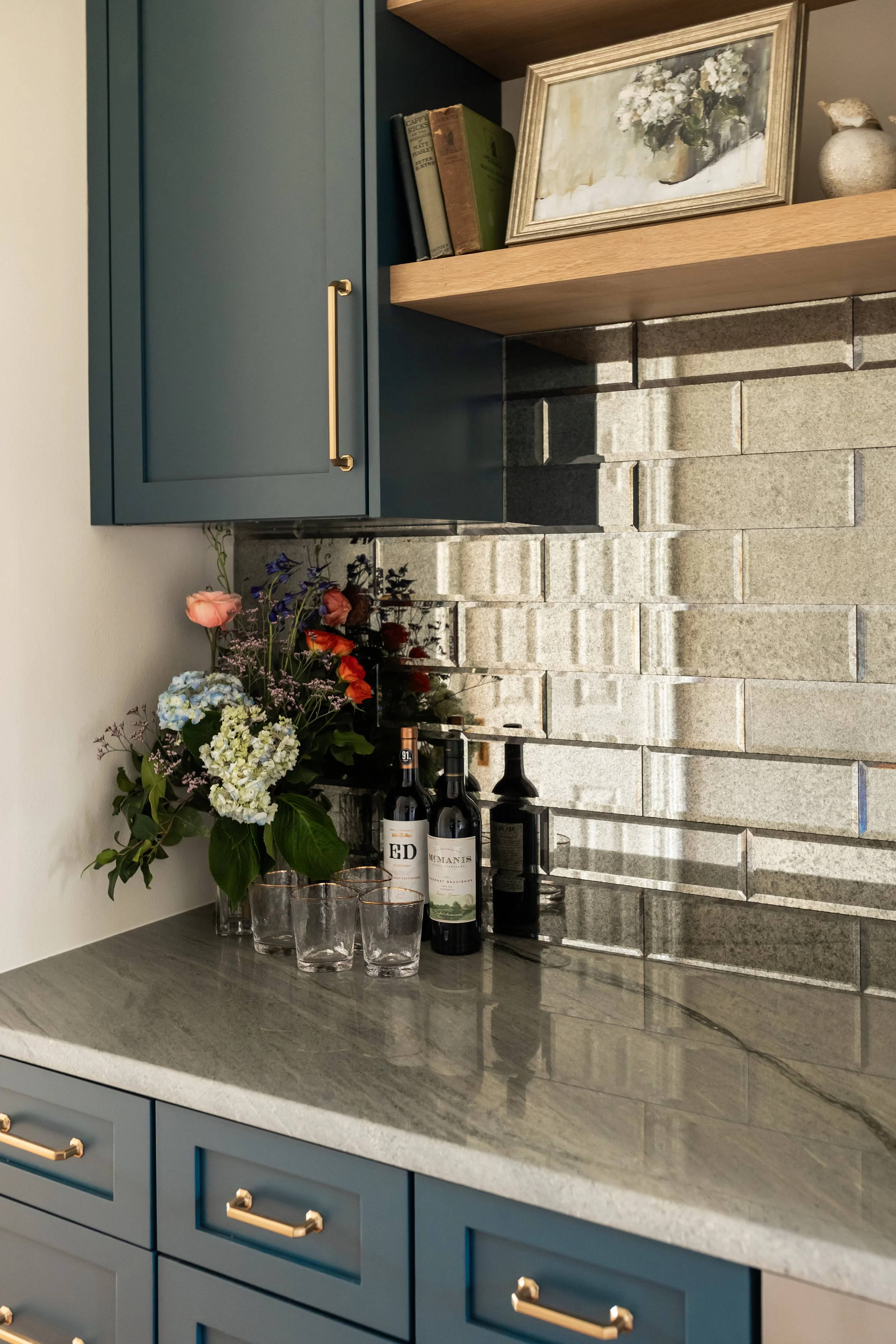 This custom bar creates a moody yet refined moment, anchored by deep blue shaker cabinetry and warm brass hardware that add richness and contrast. A dimensional stone tile backsplash brings texture and depth, while the open wood shelf softens the pal