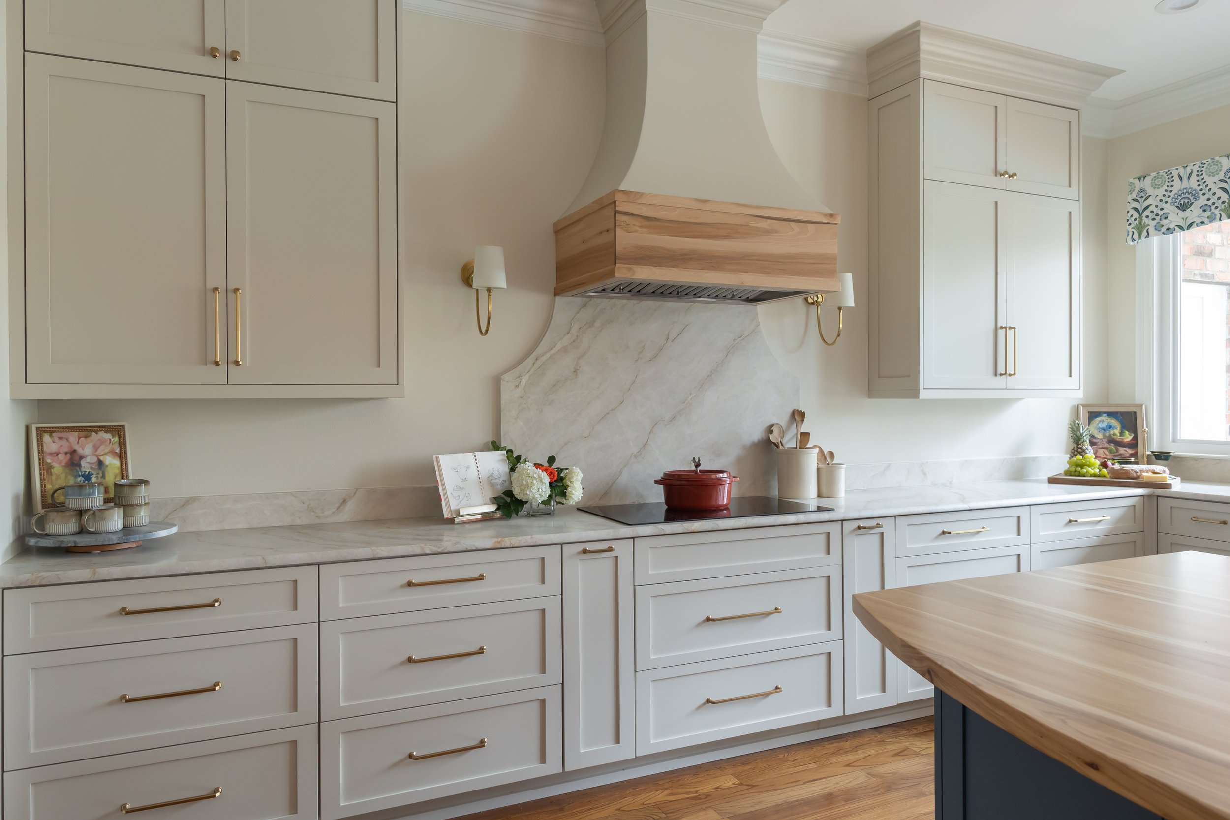The kitchen stove is centered beneath a custom wood range hood, creating a natural focal point along the back wall. A marble backsplash with soft gray veining adds elegance and contrast while keeping the palette refined. Shaker-style kitchen cabinets