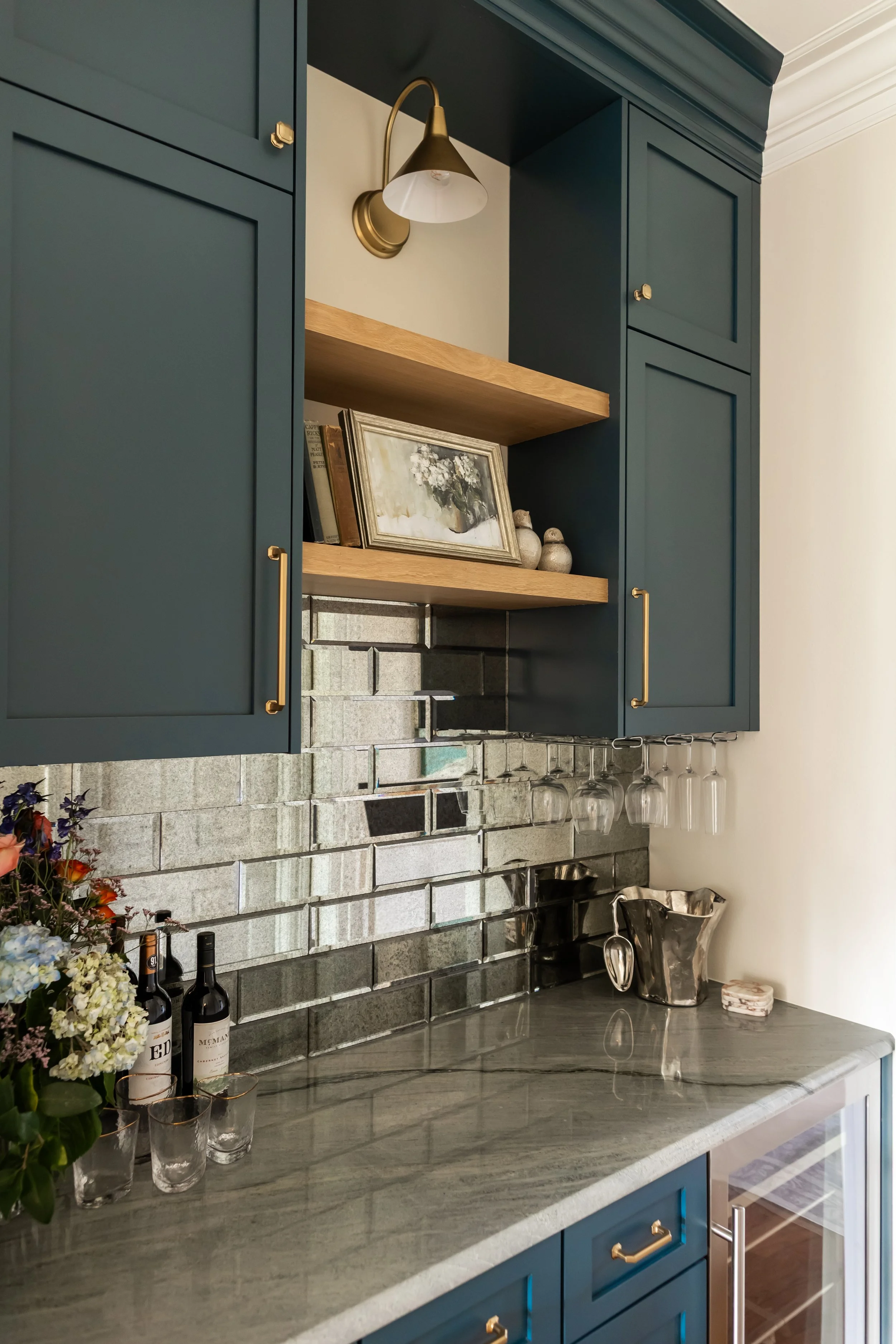 This custom bar creates a moody yet refined moment, anchored by deep blue shaker cabinetry and warm brass hardware that add richness and contrast. A dimensional stone tile backsplash brings texture and depth, while the open wood shelf softens the pal