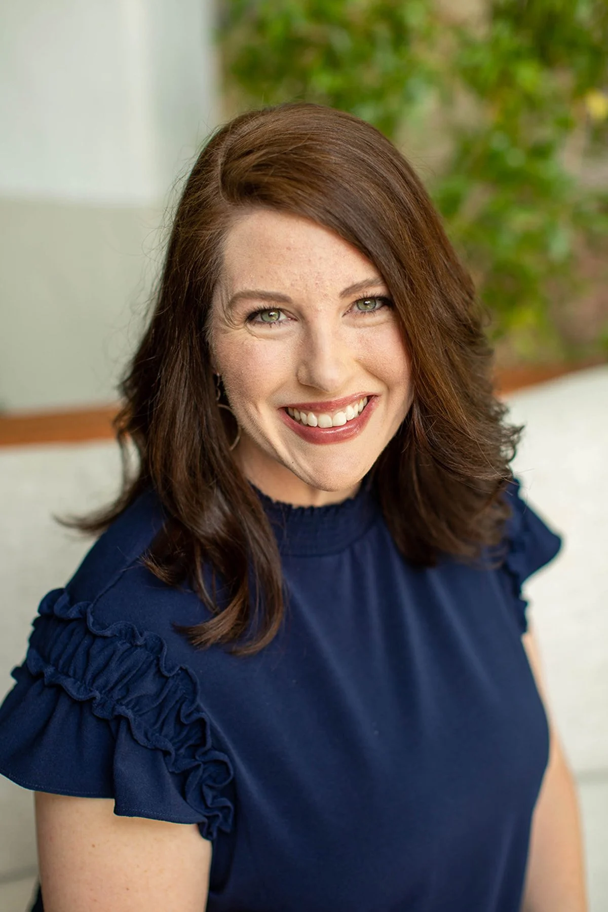 City Mom Media co-founder Jenny-Lyn Yoon wearing a navy blue top with ruffled sleeves, sitting indoors with a blurred green outdoor background.