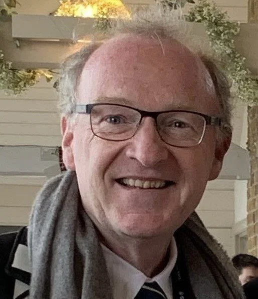 A smiling man wearing glasses, a scarf, and a suit jacket, standing outside with a house and trees in the background.