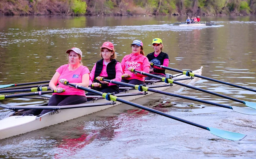 Women in a competitive rowing boat on a river, wearing pink and black athletic clothing.