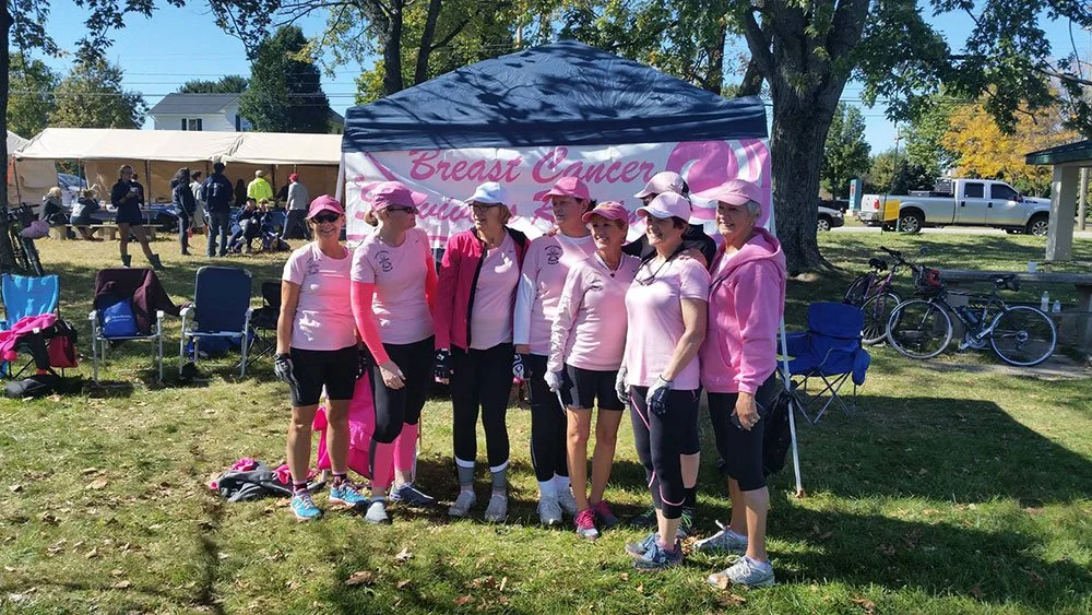 Group of women in pink clothing standing in front of a tent with a banner that reads "Breast Cancer" during a community event celebrating Breast Cancer awareness.