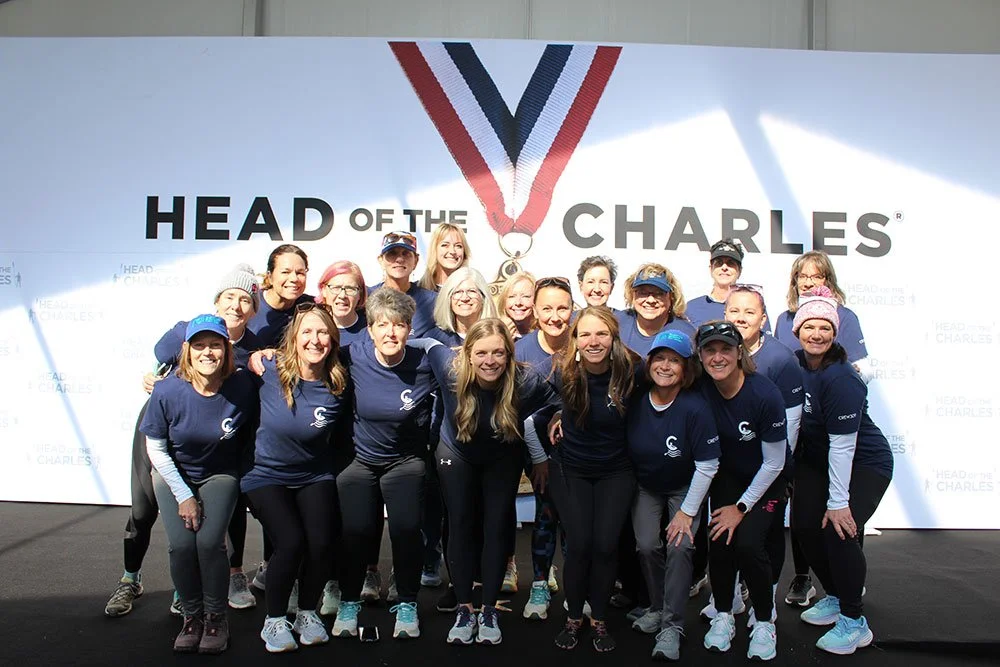 Group of women in matching navy blue shirts and athletic wear, standing in front of a large backdrop with the words 'HEAD OF THE CHARLES' and a medal symbol, smiling for a photo.