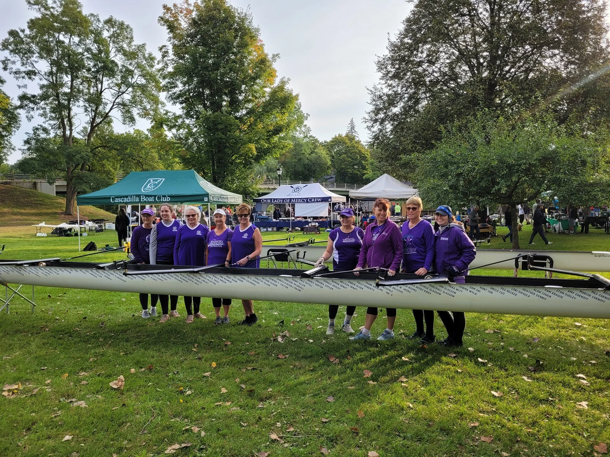 Group of women in purple and black athletic clothing standing behind a rowing shell on a grassy field at a outdoor event with tents and trees in the background.