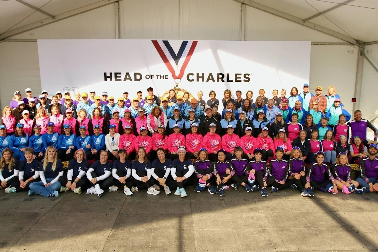 Group photo of women and girls in colorful athletic clothing under a tent with a large sign that reads "Head of the Charles," featuring a medal with a ribbon. They are smiling and posing for the picture.