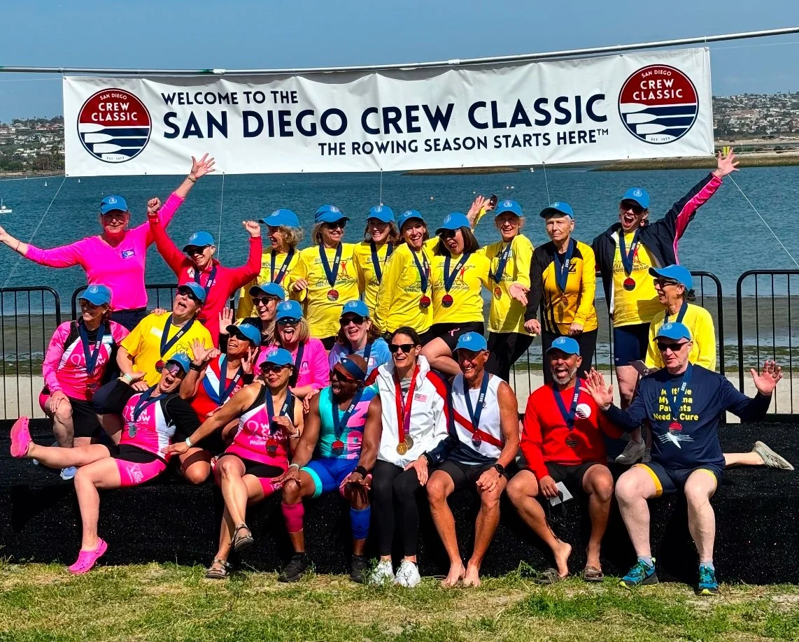 Group of rowers and coaches posing with medals in front of a banner that reads "Welcome to the San Diego Crew Classic, the rowing season starts here." They are wearing colorful athletic clothing and hats, standing near a body of water.