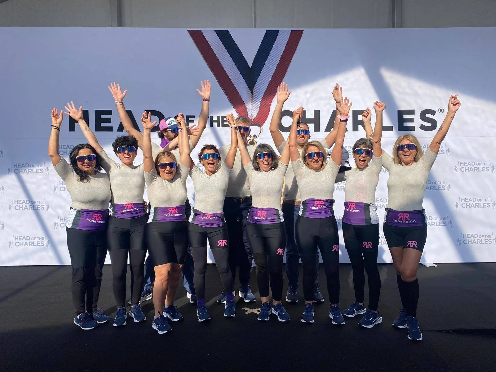Group of women standing in front of a backdrop with a medal graphic, raising their arms in celebration, all wearing matching gray shirts, black pants, blue sunglasses, and purple waistbands with pink ribbons.