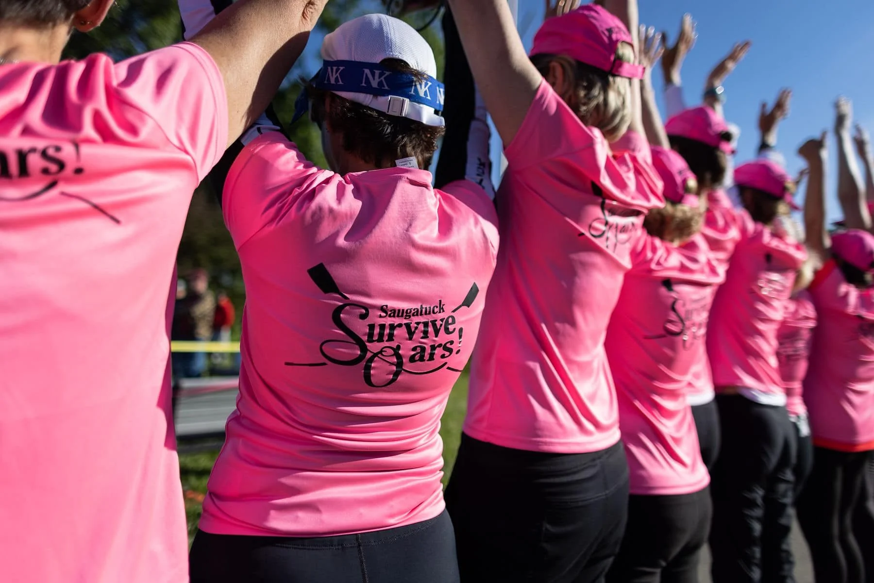 Group of women wearing pink shirts with 'Saugatuck Survive Oars!' logo, participating in a team rowing event outdoors on a sunny day.