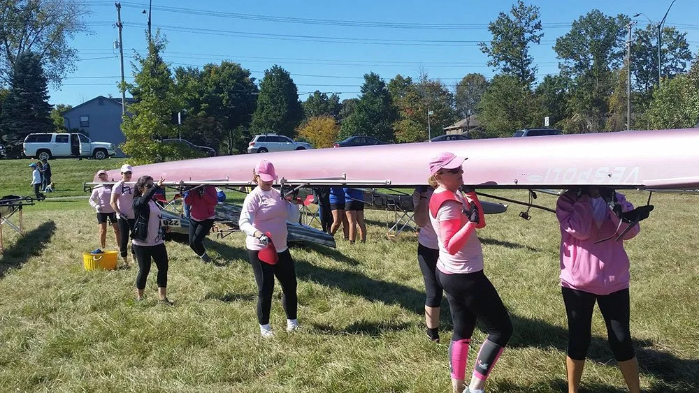A group of women in athletic clothing and pink hats standing on grass, carrying a large pink rowing shell.
