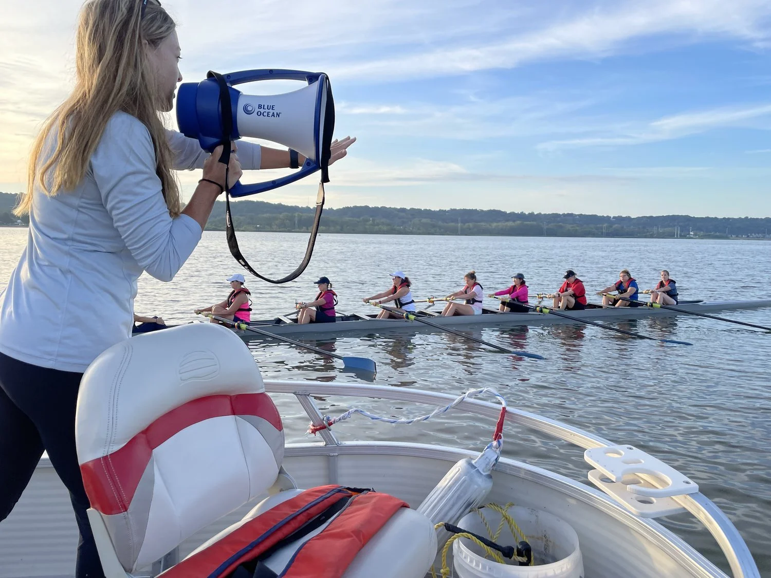 A woman with long blonde hair holding a megaphone on a boat, coaching a rowing team on a lake at sunset. Eight rowers in matching pink and white uniforms are rowing in a long boat.