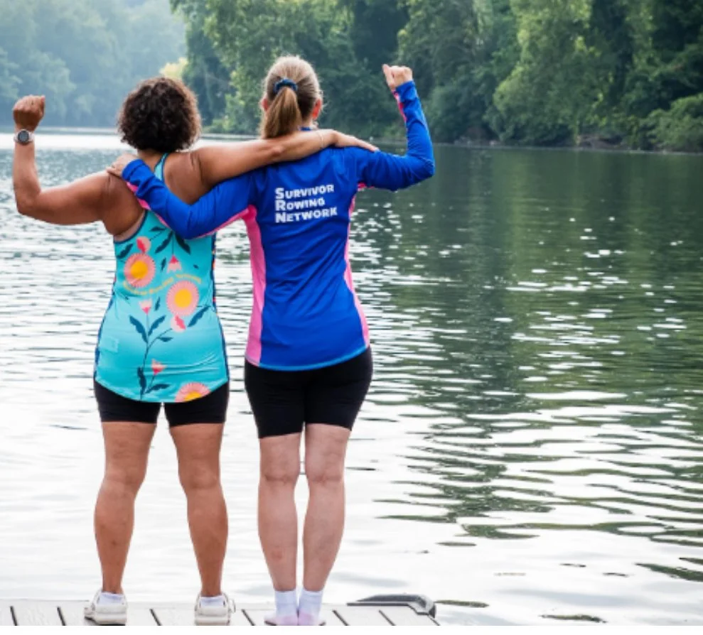 Two women with their backs to the camera, standing on a dock by a river, embracing and flexing their biceps. One woman wears a colorful floral tank top, and the other wears a blue jacket that says 'Survivor Rowing Network.' The scene is outdoors with