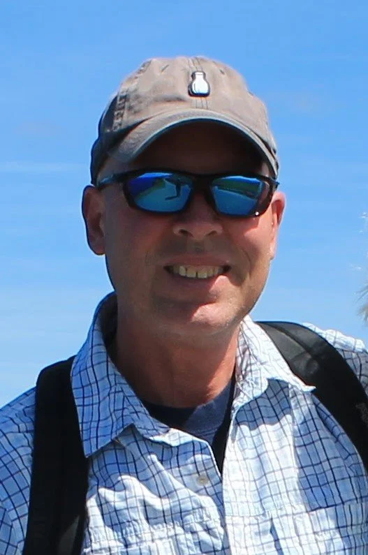 Man wearing a beige hat, sunglasses, and a plaid shirt outdoors under a blue sky.