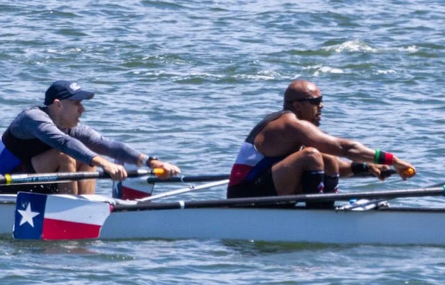 Two men rowing a boat on the water, with one wearing sunglasses and another wearing a cap.