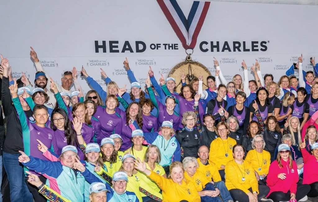 Group of people celebrating at the Head of the Charles rowing event, with medals and event merchandise, large medal hanging in the background.