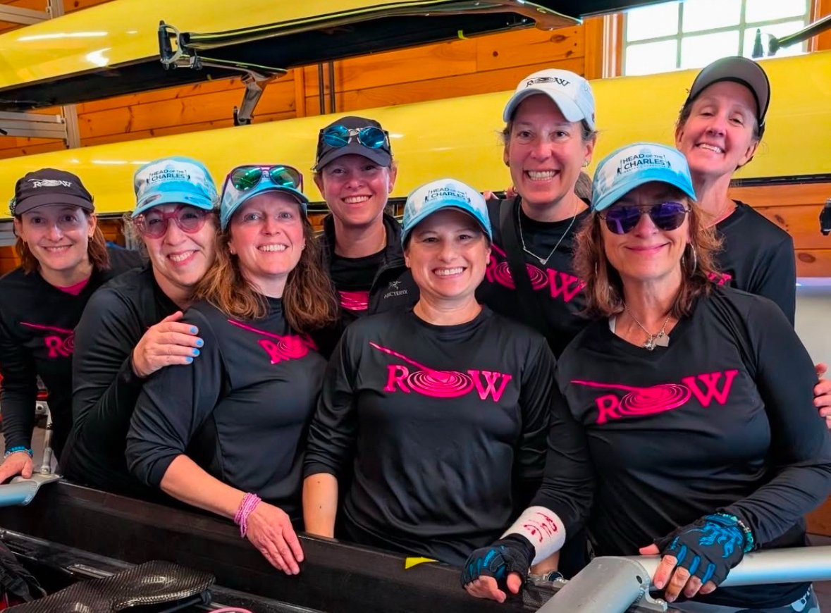 Group of women in black and pink matching shirts with 'ROW' logo, posing together in a boat storage area with yellow kayaks and wooden walls behind them. Some wearing caps and sunglasses, smiling at the camera.