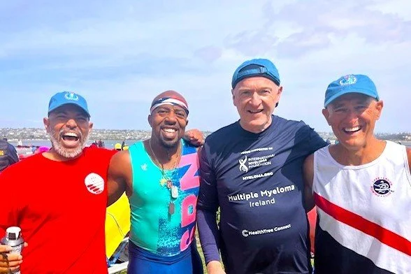 Four people smiling and posing for a photo outdoors during a race or event, with water and buildings in the background.