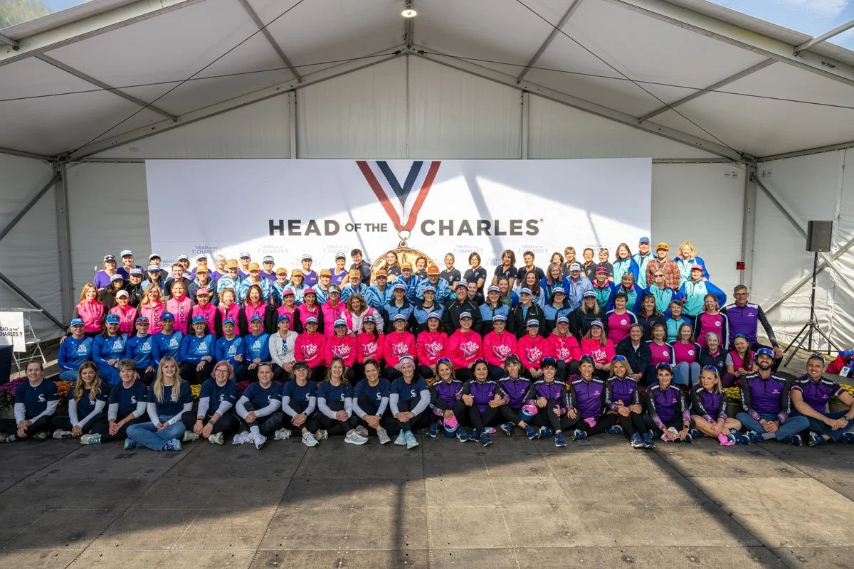 Group of people posing on stage at the Head of the Charles rowing event, with a large sign behind them displaying the event name.
