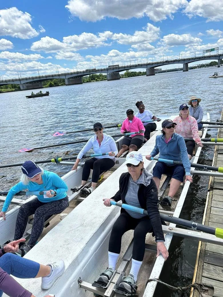Group of women in a rowing boat on a river, preparing to row, under a partly cloudy sky, with a bridge in the background.