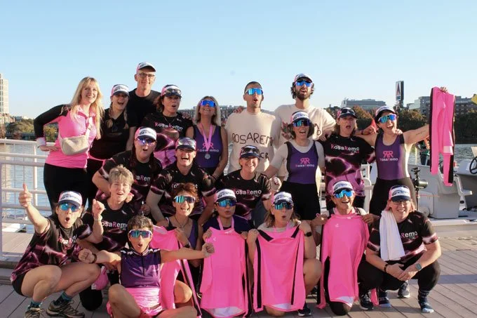 Group of people in athletic gear on a boat deck, posing for a photo with some holding pink sports jerseys, wearing sunglasses and caps, under a clear blue sky.
