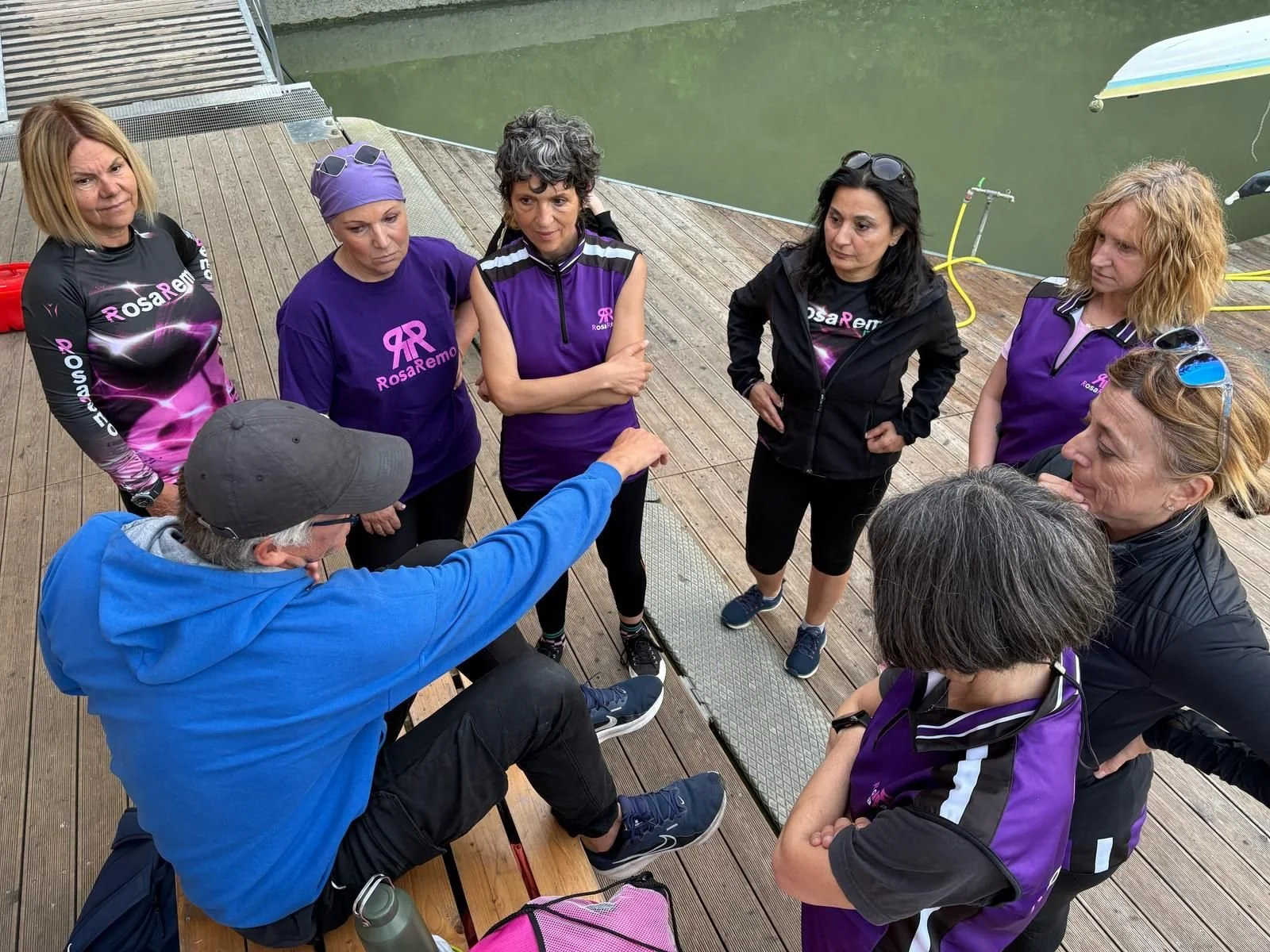 Group of women in matching purple and black shirts listening to a man in a blue hoodie who is sitting on a bench and pointing.