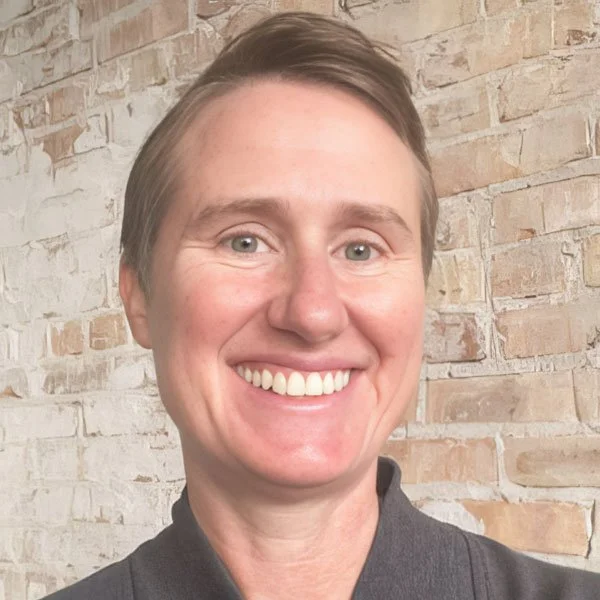 A person with short brown hair, smiling, wearing a dark shirt, standing in front of a brick wall.