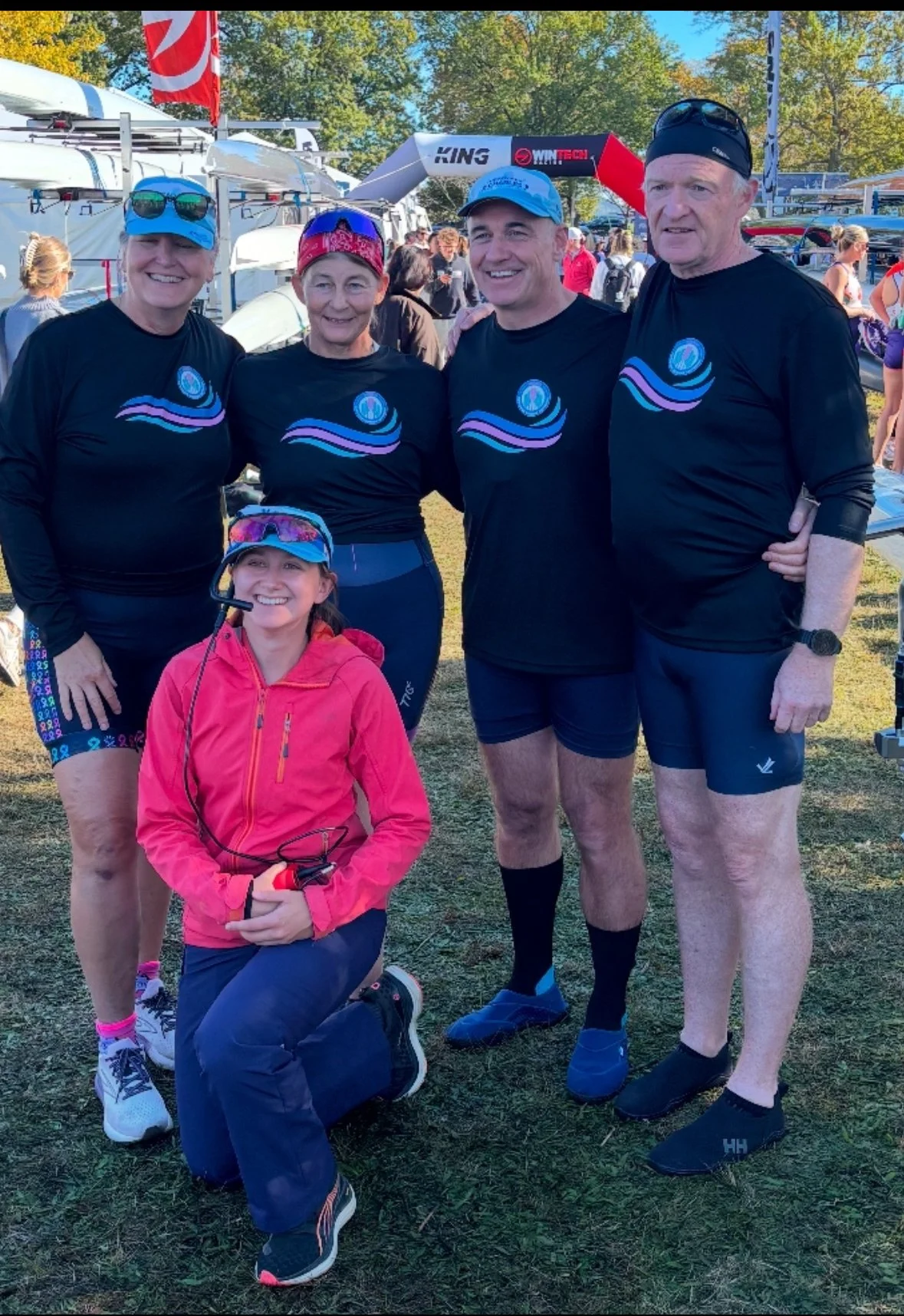 A group of five people, four adults and one child, dressed in athletic clothing, standing outdoors at a sporting event. They are smiling at the camera, wearing matching black shirts with a blue and purple logo, possibly a swim team or athletic group.