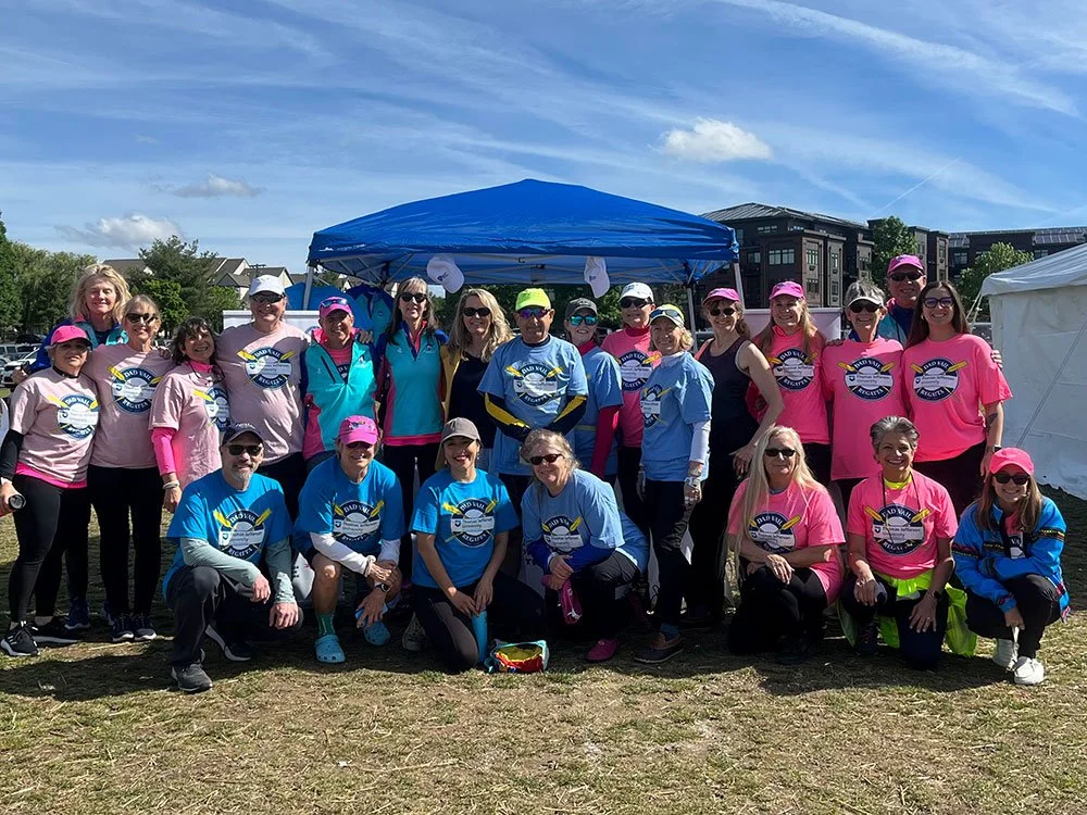 Group photo of people wearing pink and blue shirts and hats at an outdoor event, under a blue canopy on a grassy area with buildings and trees in the background.