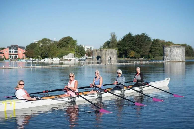 Five women rowing a boat on a calm river with trees, buildings, and a stone fortress in the background.