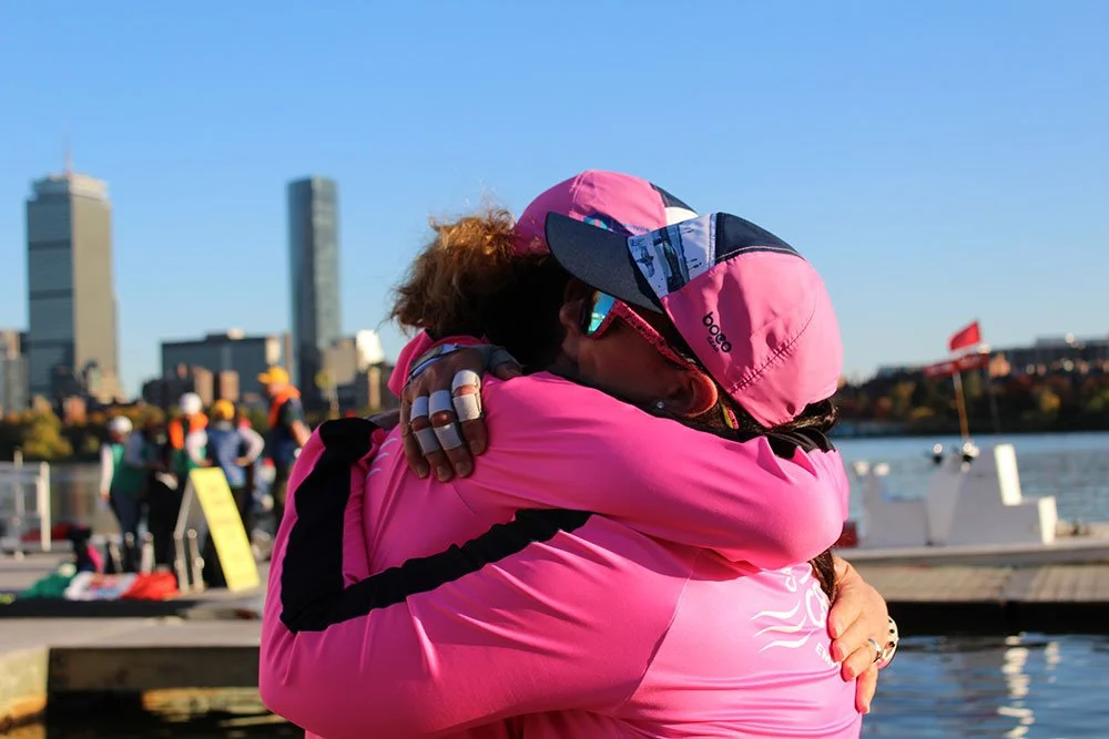 Two women hugging on a dock with a city skyline in the background; one woman wears a pink jacket and cap, the other is hidden in the embrace.
