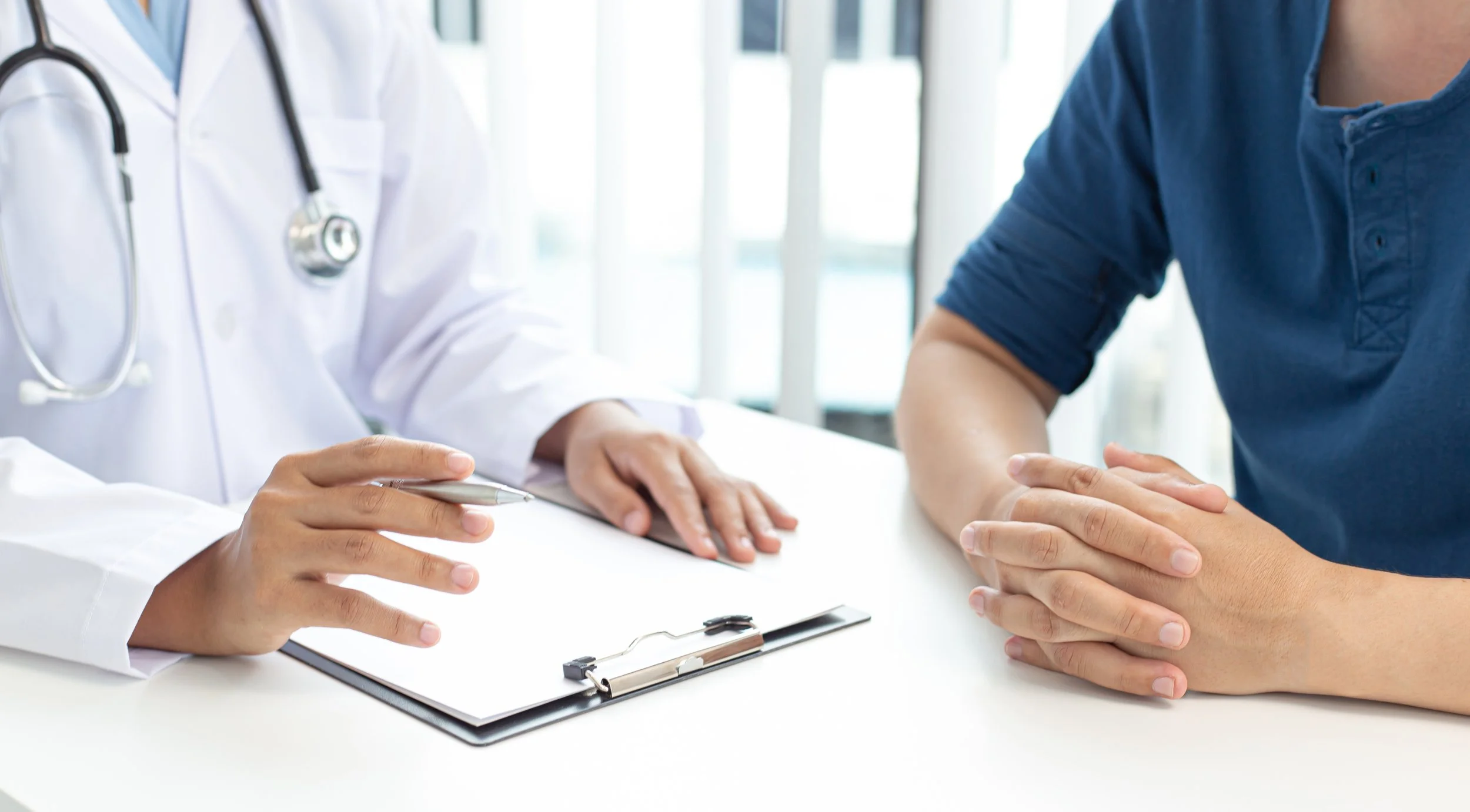 Doctor and patient in consultation, with doctor holding a pen and a clipboard on the table.