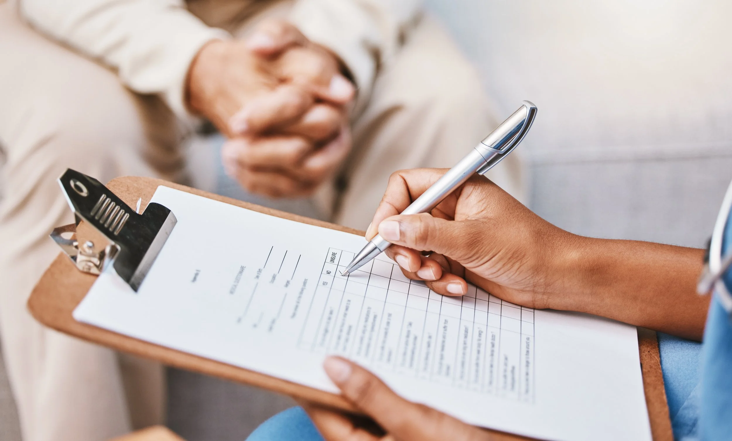 A healthcare professional is filling out a medical form on a clipboard during a consultation, with a patient sitting across and clasping their hands together.