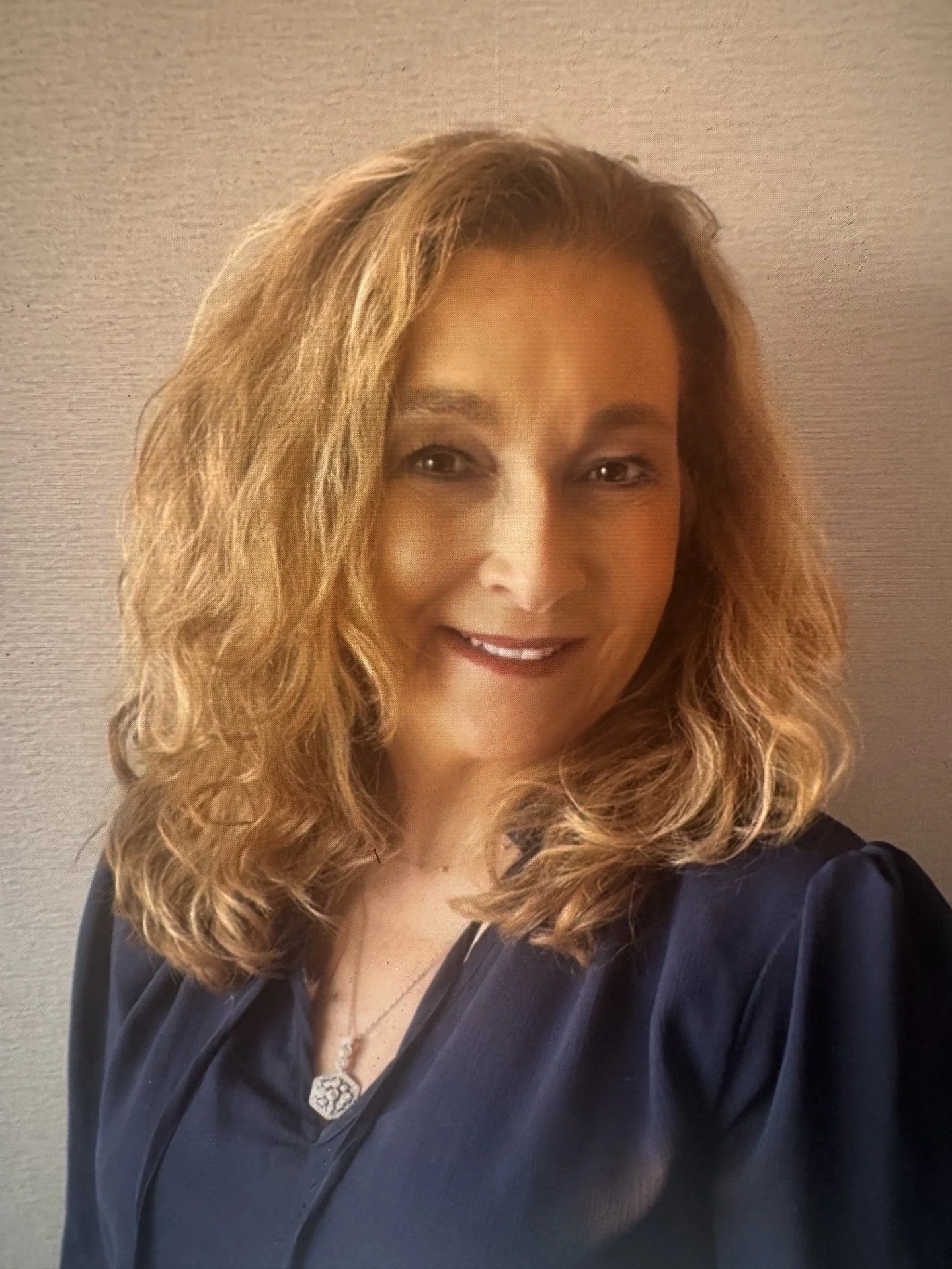 A woman with shoulder-length wavy blonde hair, smiling, wearing a dark top, standing against a neutral-colored textured wall.