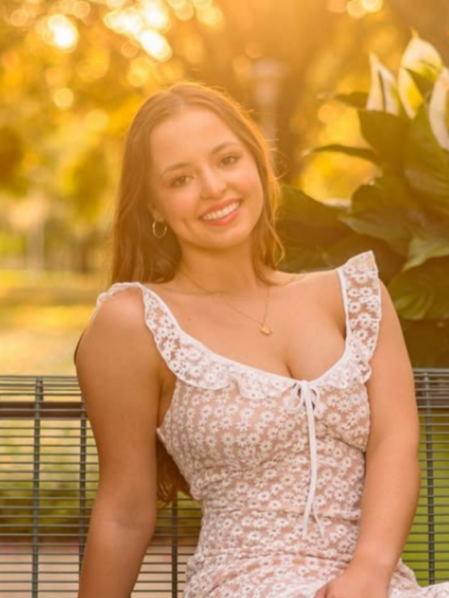 A young woman with long brown hair sitting on a park bench in warm sunlight, wearing a white floral dress with ruffled sleeves and a bow in front, smiling at the camera with trees and large white flowers in the background.