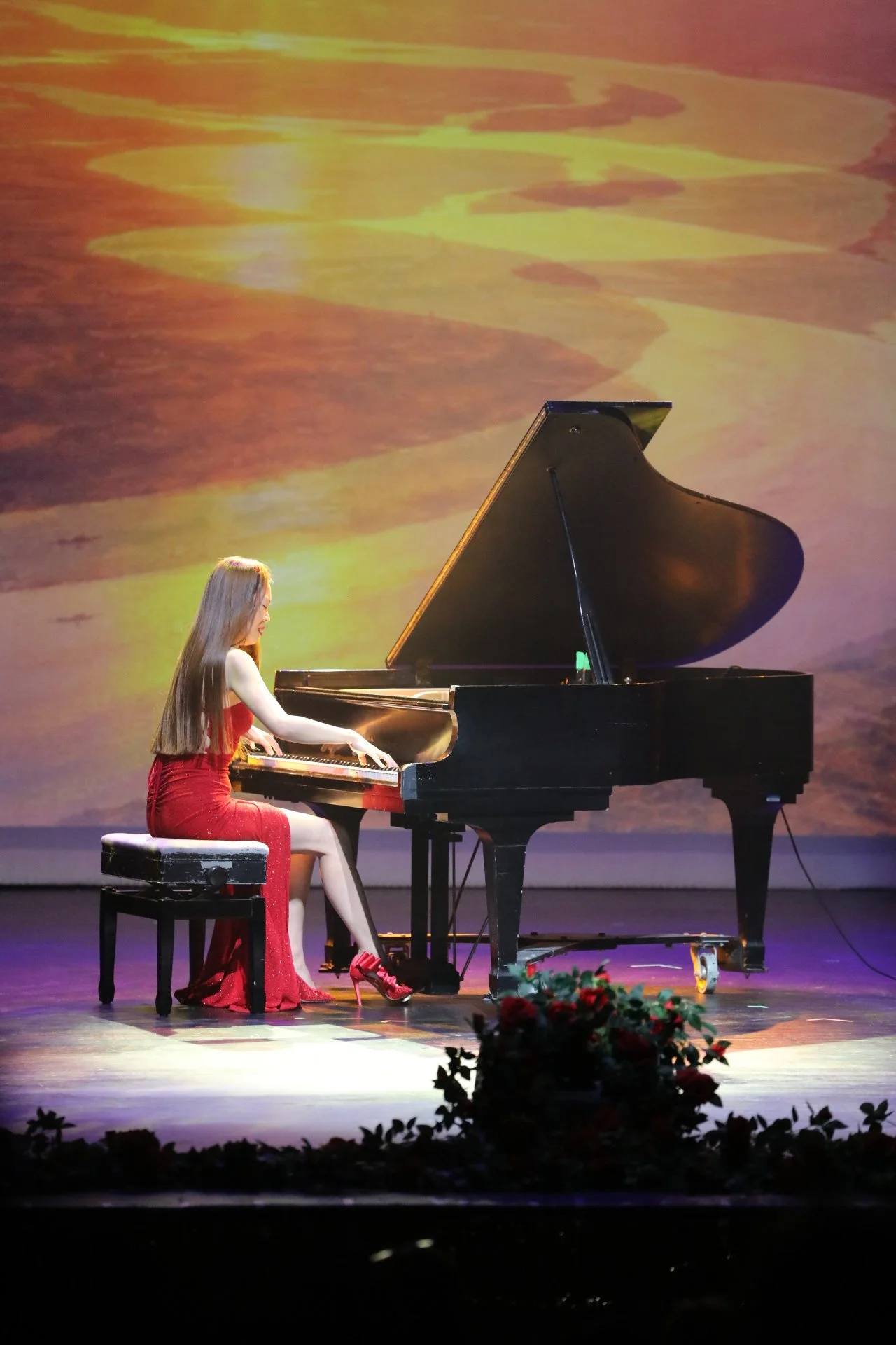 A young woman in a red dress playing a grand piano on stage with a colorful sunset background projection.