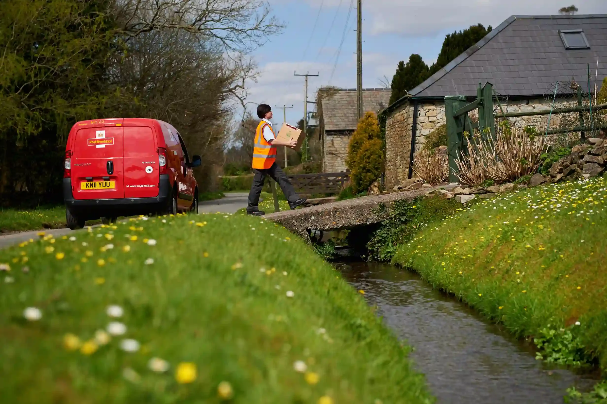 Royal Mail delivery woman carrying package to door of countryside house.