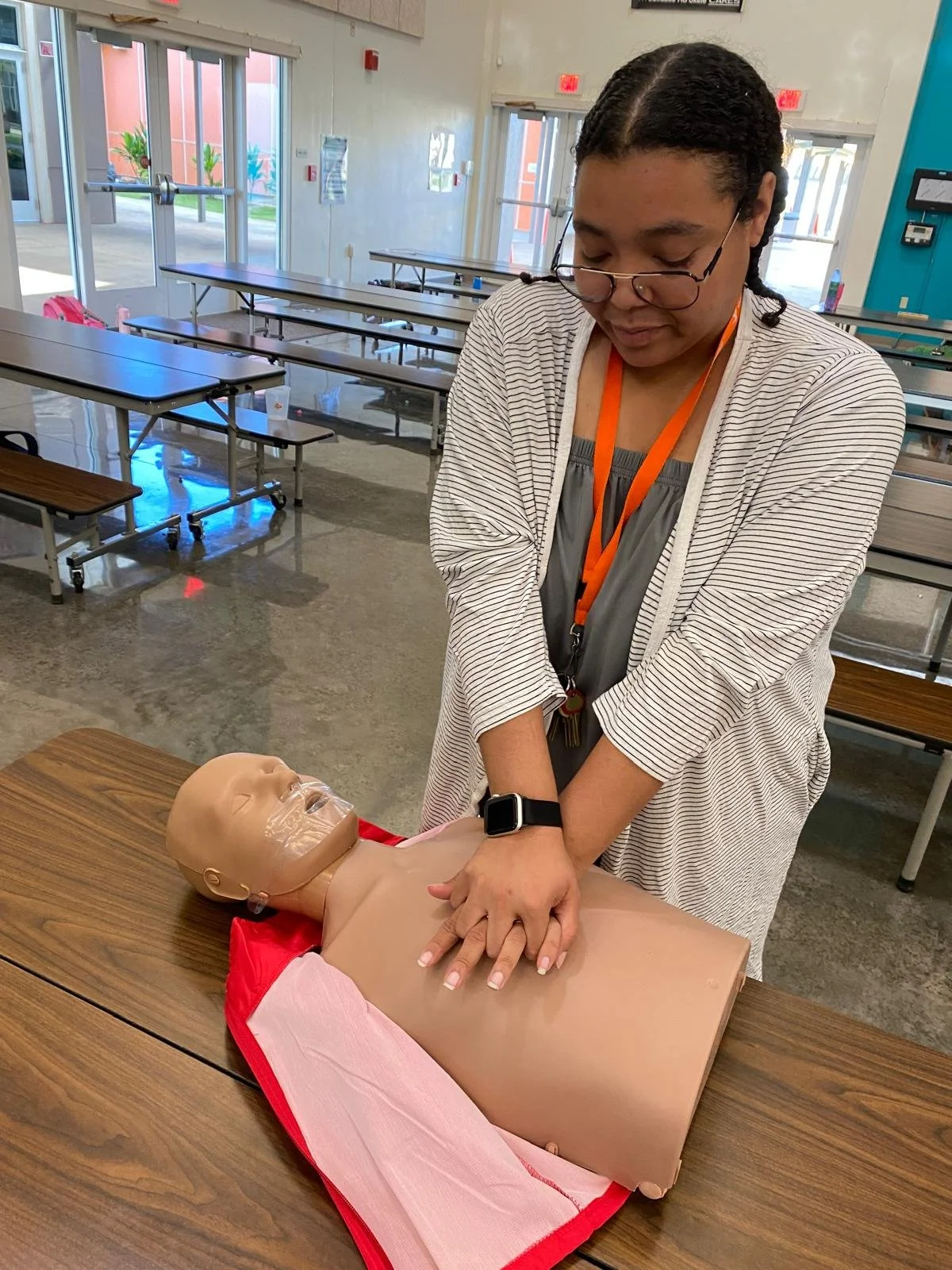 A woman practicing CPR on a medical training mannequin lying on a table inside a room with tables and chairs.