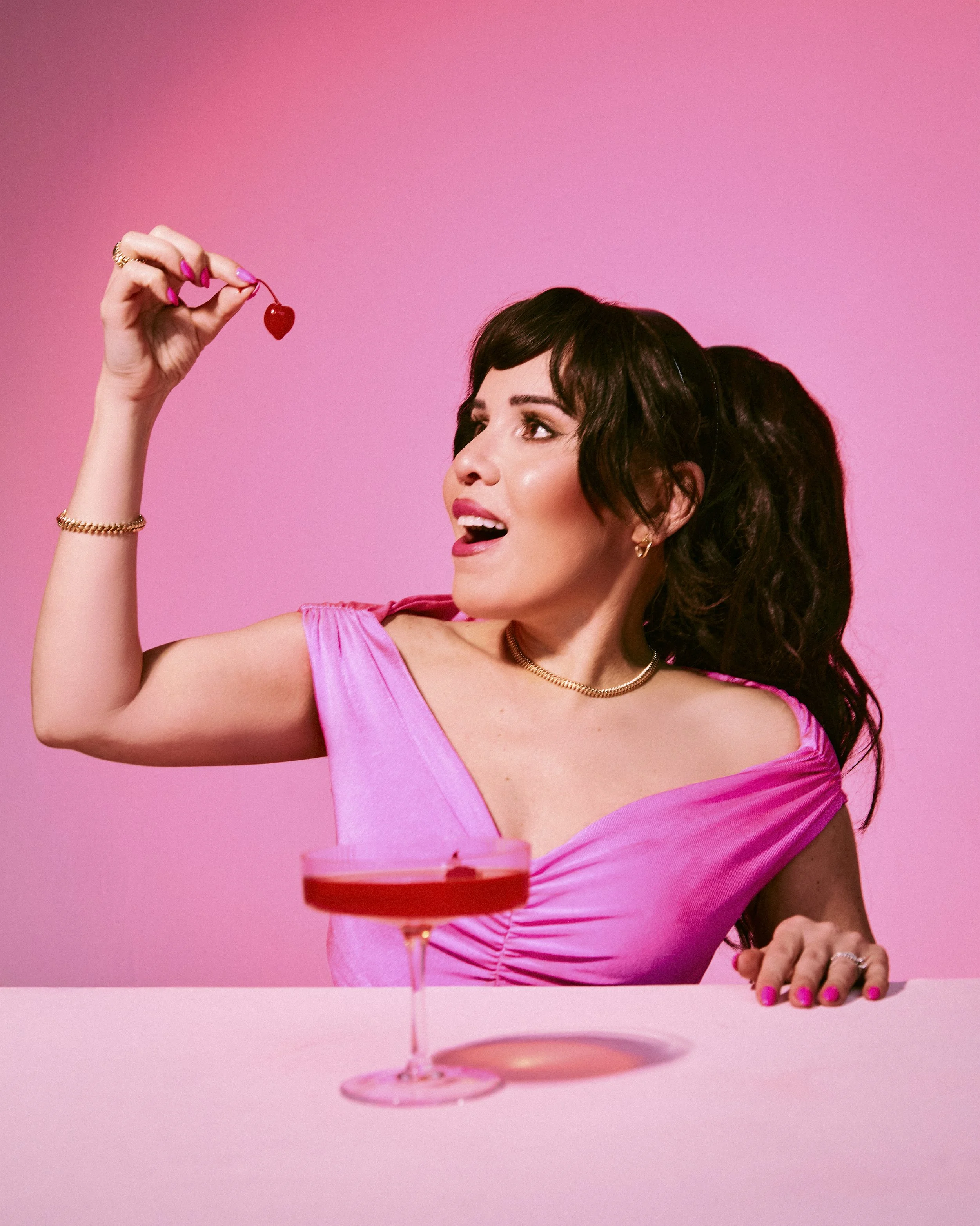 A woman with dark hair and pink nails holding a red cherry above her open mouth, with a pink background and a pink cocktail glass in the foreground.
