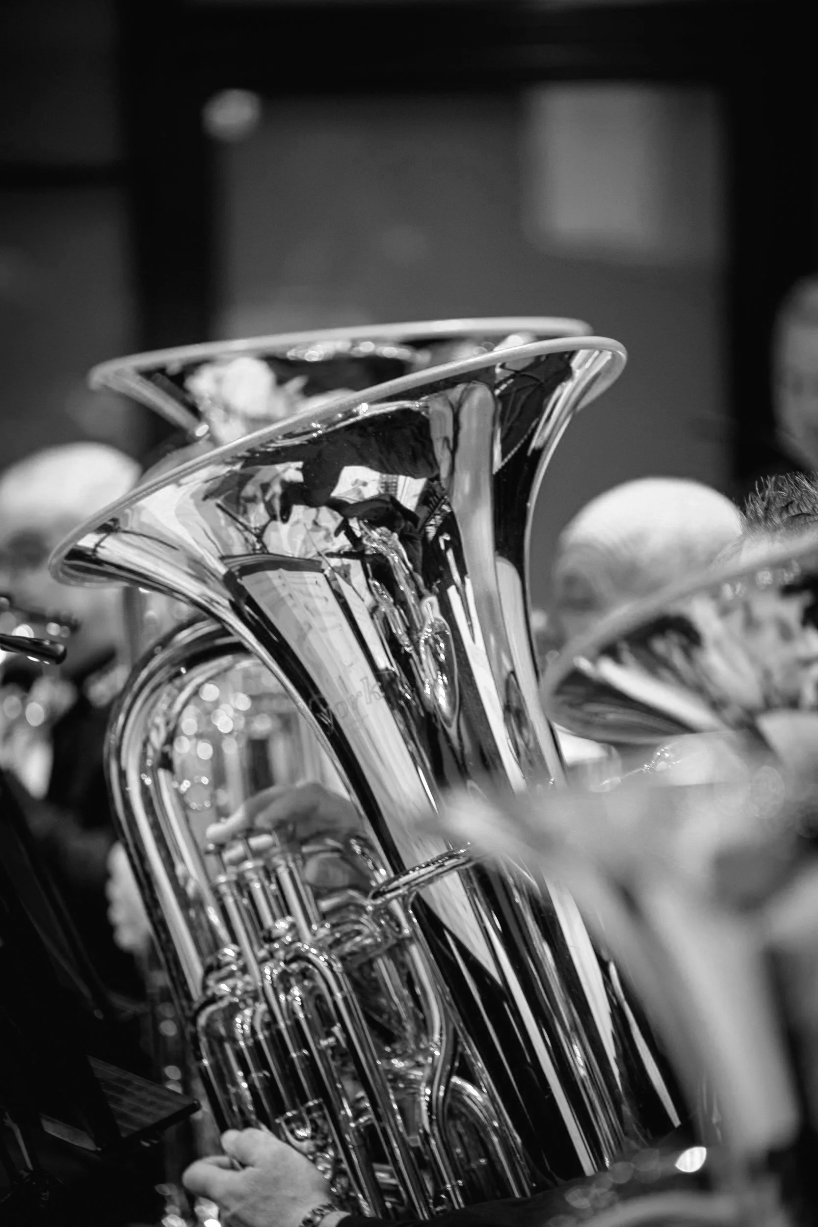 Close-up of a shiny, reflective tuba among other brass instruments, with people wearing hats in the background.