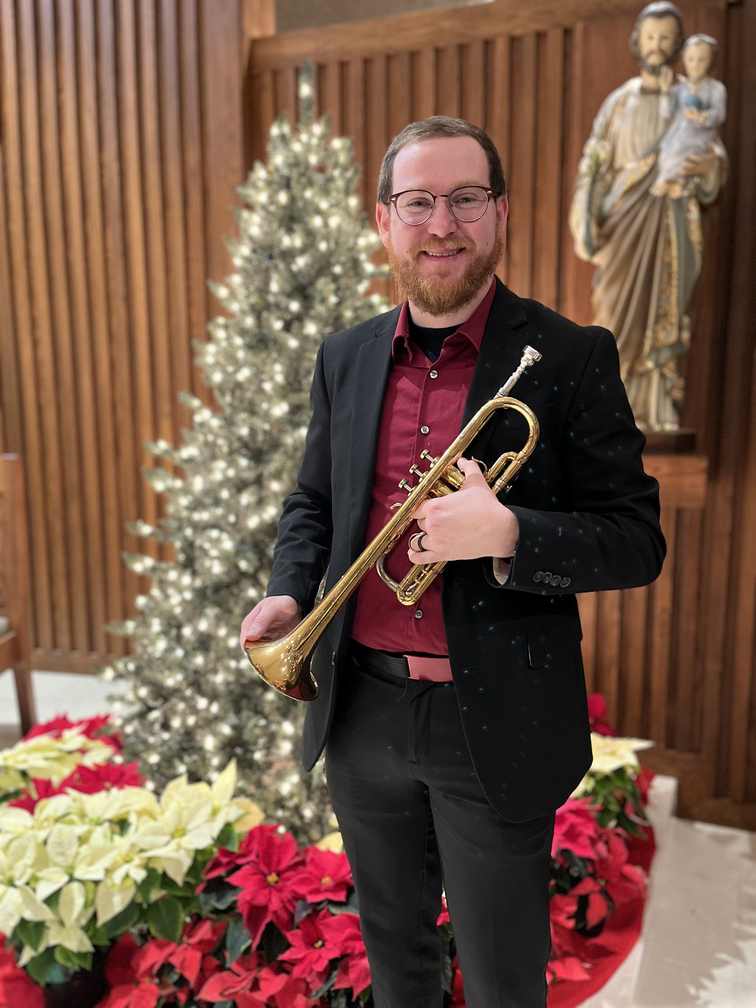 A man with glasses and a beard holding a trumpet, dressed in a black blazer and red shirt, standing in front of a decorated Christmas tree and poinsettias, with a nativity statue in the background.