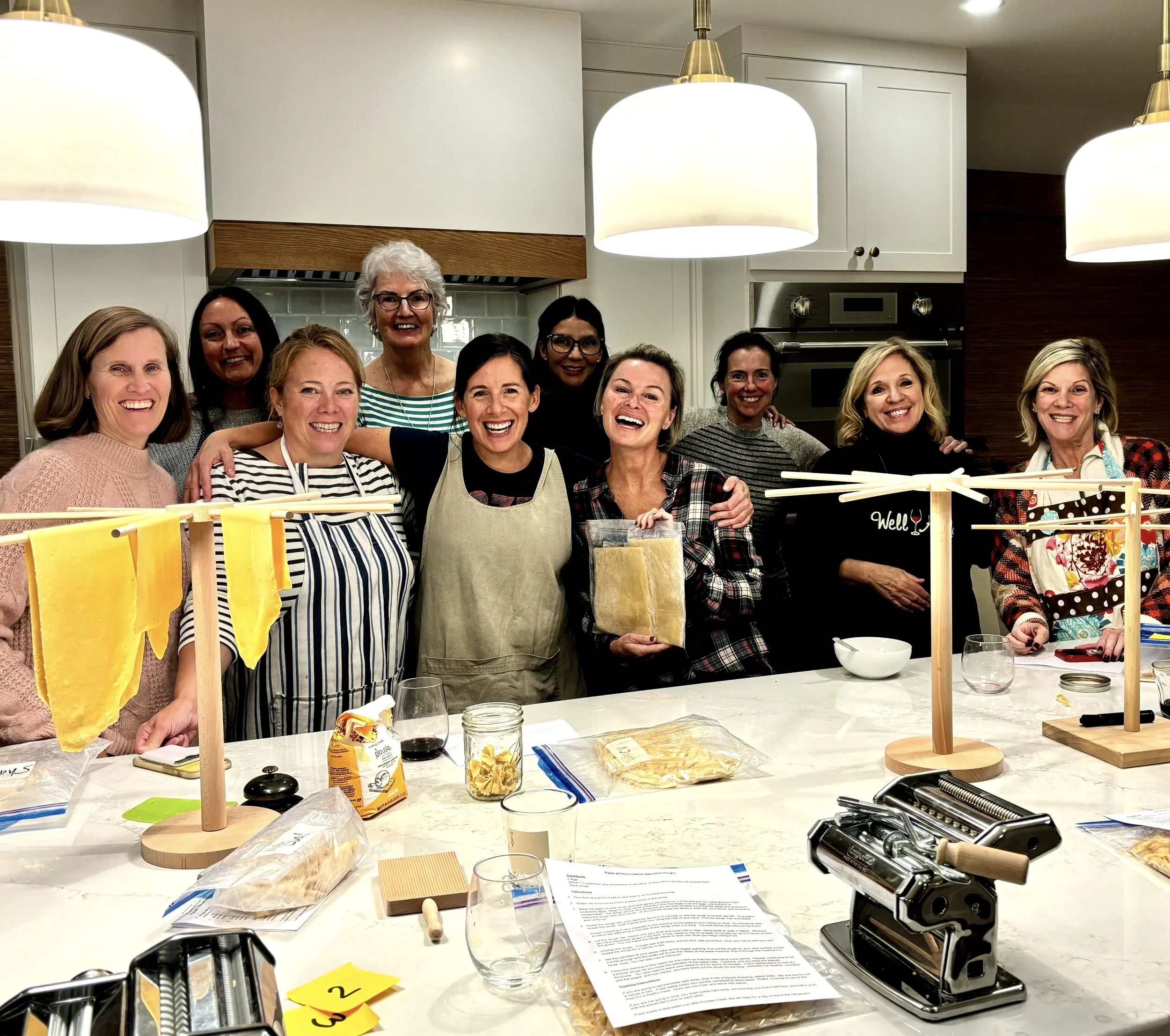 Group of smiling women gathered around a kitchen island with pasta-making equipment and ingredients, celebrating a cooking class or event.