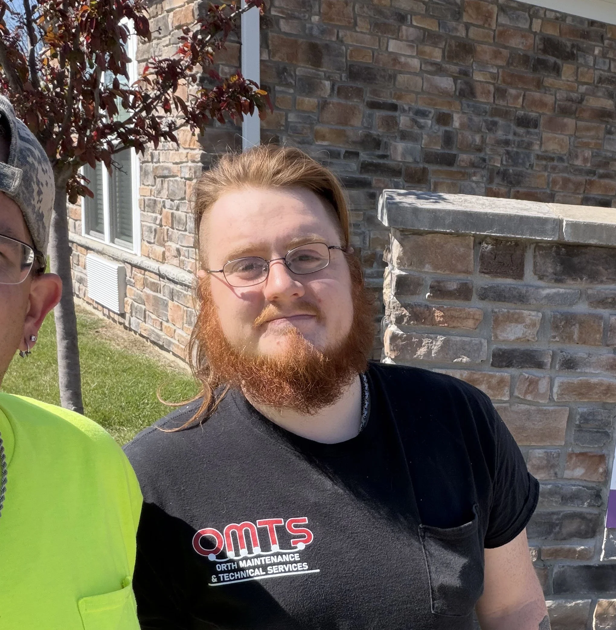 A young man with glasses and brown hair wearing a neon yellow shirt standing in front of a stone wall and a sign that says 'Otto'.