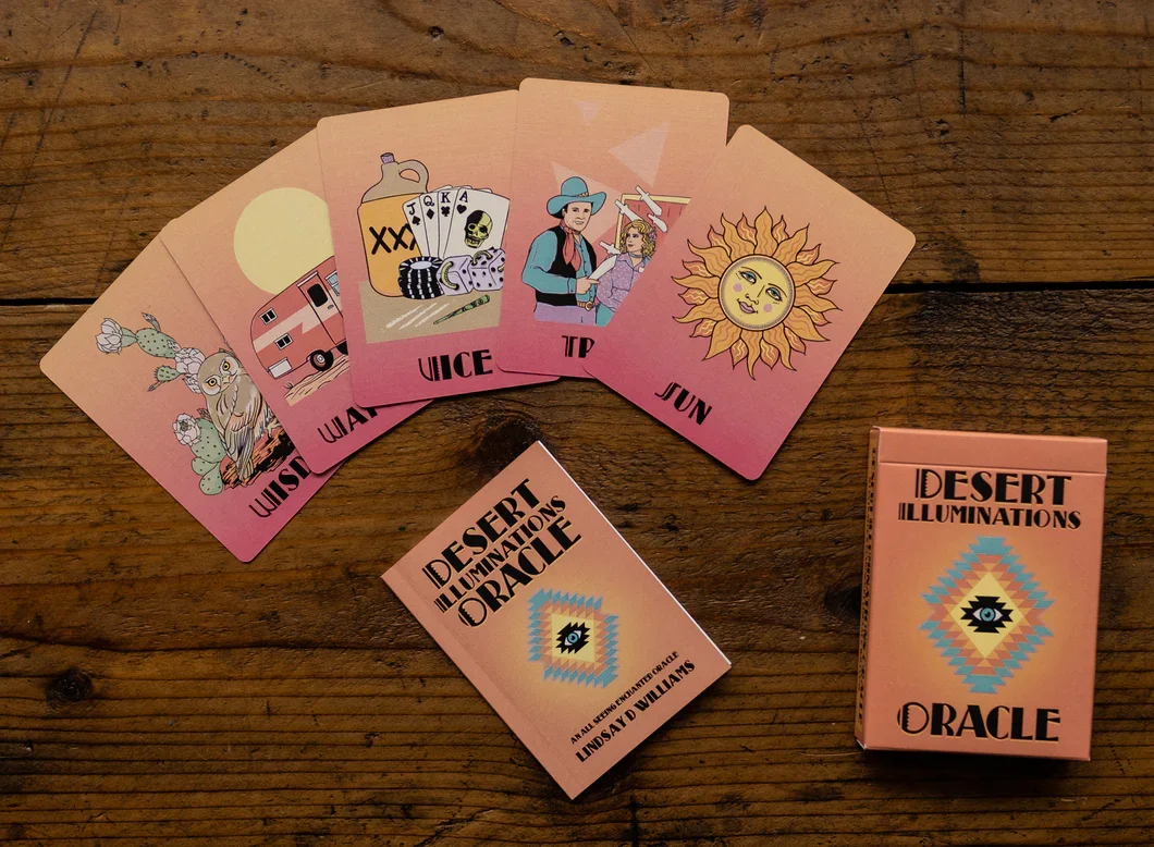 Set of five illustrated playing cards with pink backgrounds, depicting a cactus, a camper trailer, whiskey bottles with cards, a woman with a hat, and a sun with a face, arranged on a wooden surface. A deck box titled 'Desert Illuminations Oracle' with a geometric eye design is also on the surface.
