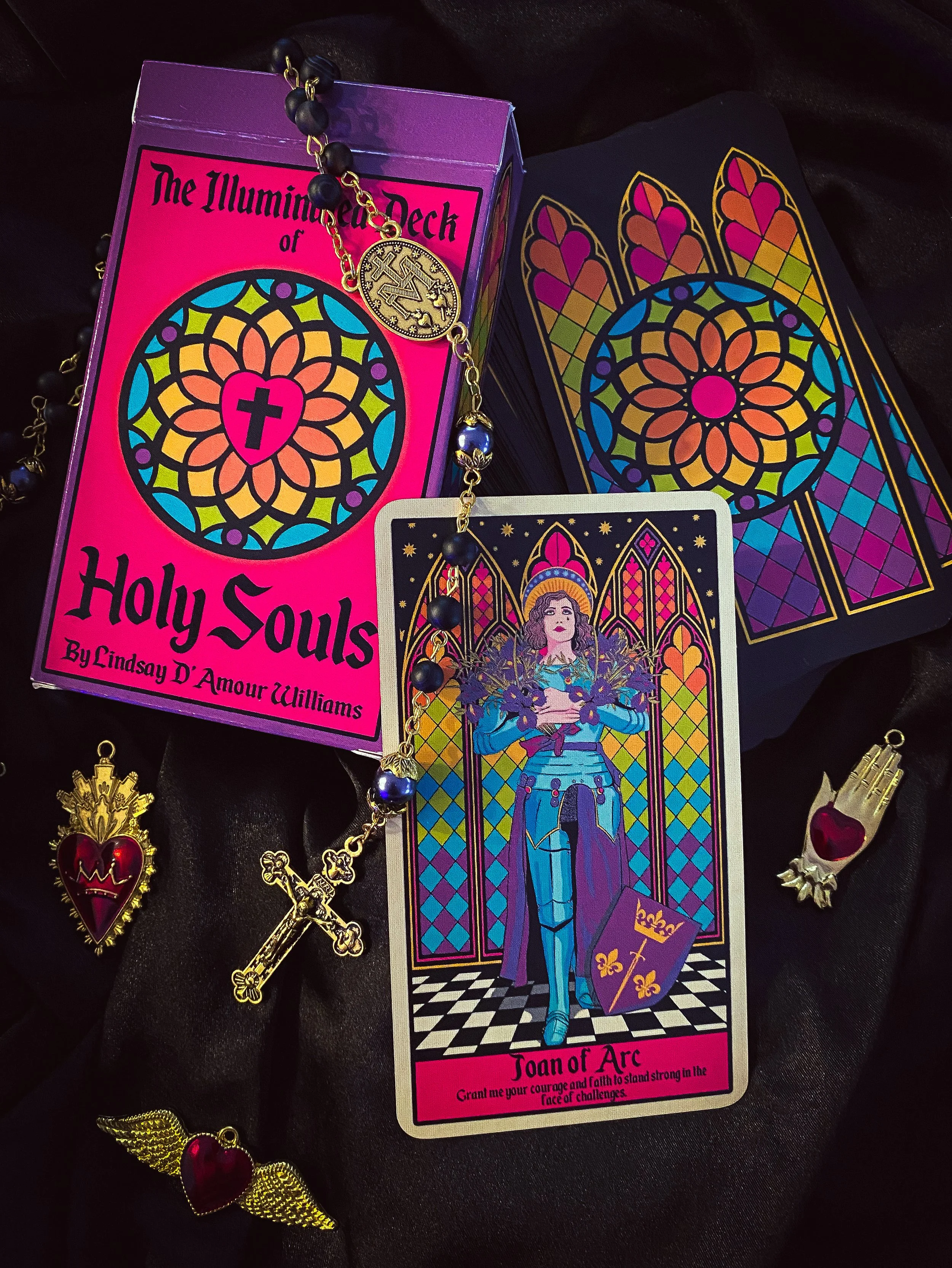 A collection of Tarot and spiritual themed items: a pink tarot card box labeled 'The Illuminated Deck of Holy Souls' with a colorful stained-glass window design, a tarot card titled 'Joan of Arc' with an illustration of Joan in armor holding a sword and shield, and various jewelry pieces including a rosary with a cross, a heart with wings, a hand with a heart, and a heart with wings.