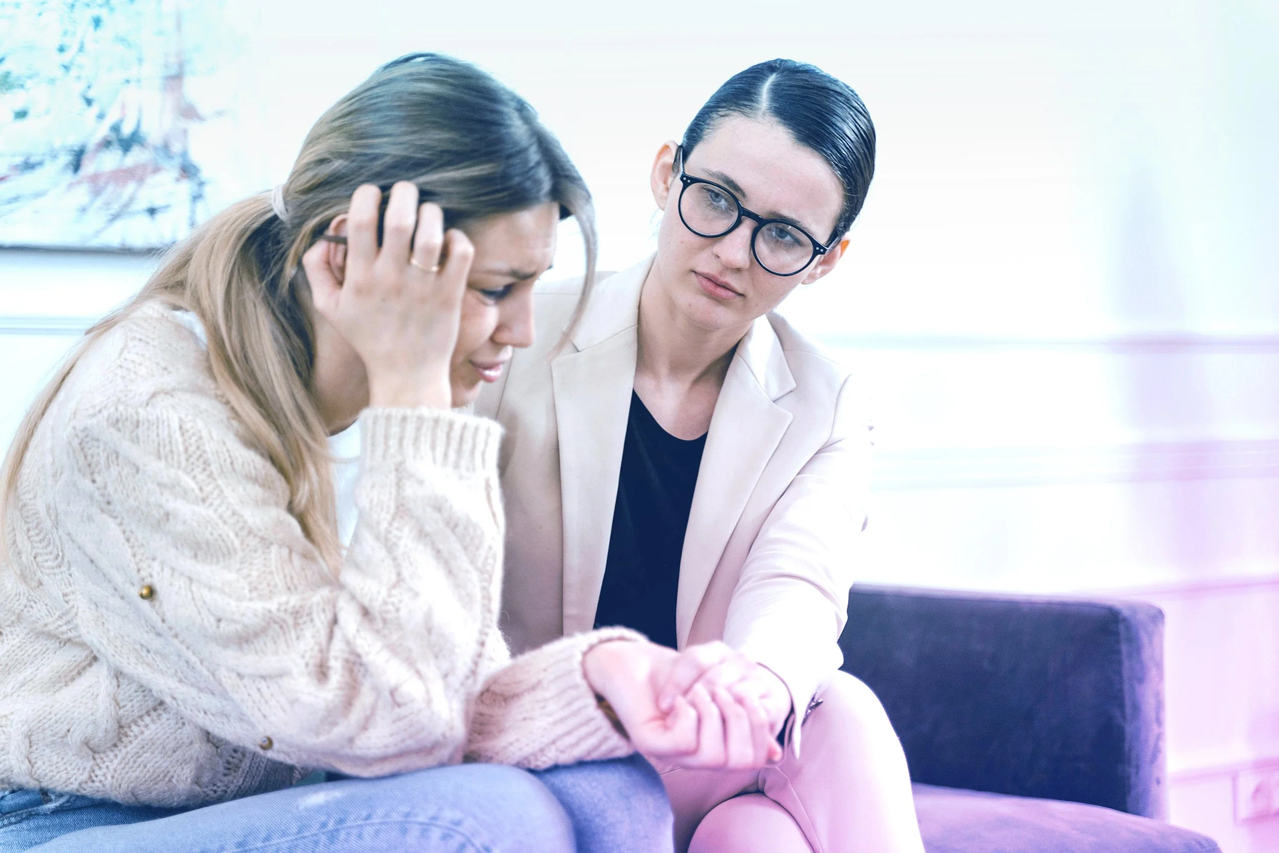 A woman is holding her head and crying, while another woman sits beside her, comforting her with a hand on her arm, in a bright room.